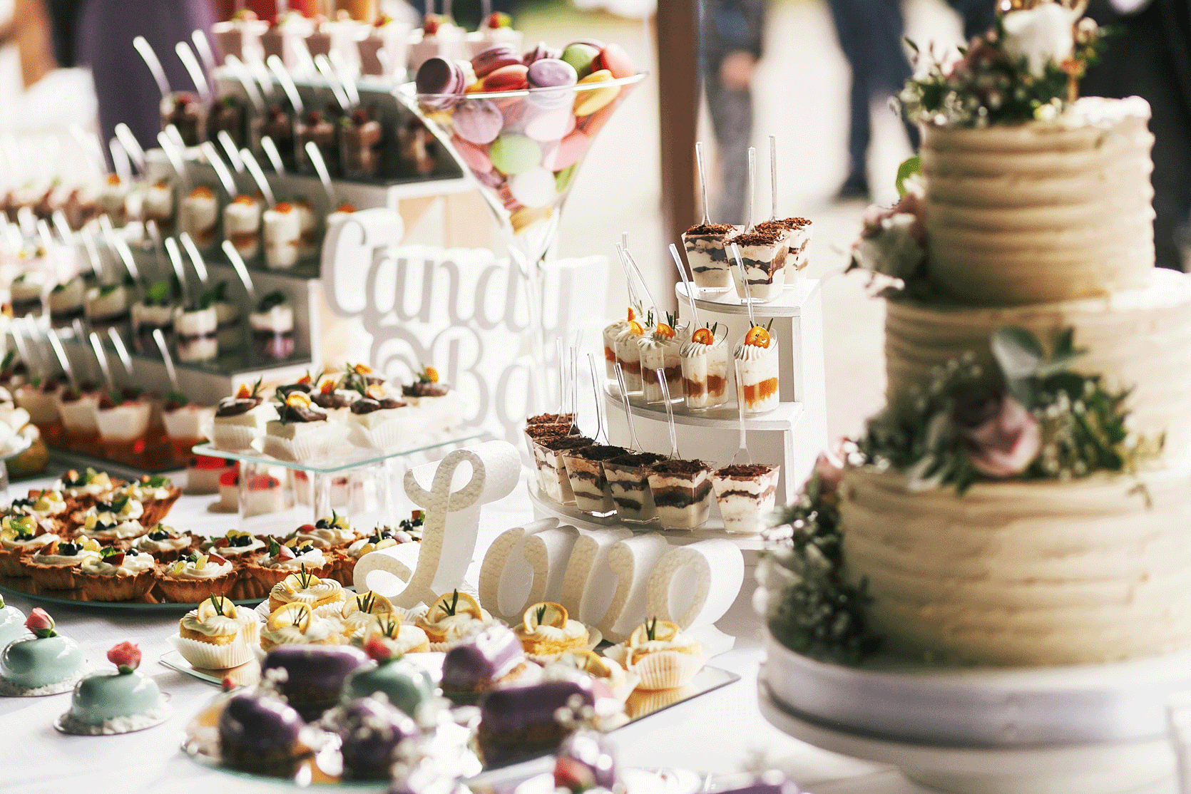 Wedding dessert table with various cakes, cupcakes, and parfaits, including a large three-tiered wedding cake with floral decorations.