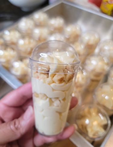 A clear plastic cup filled with vanilla pudding and topped with crushed cookies.