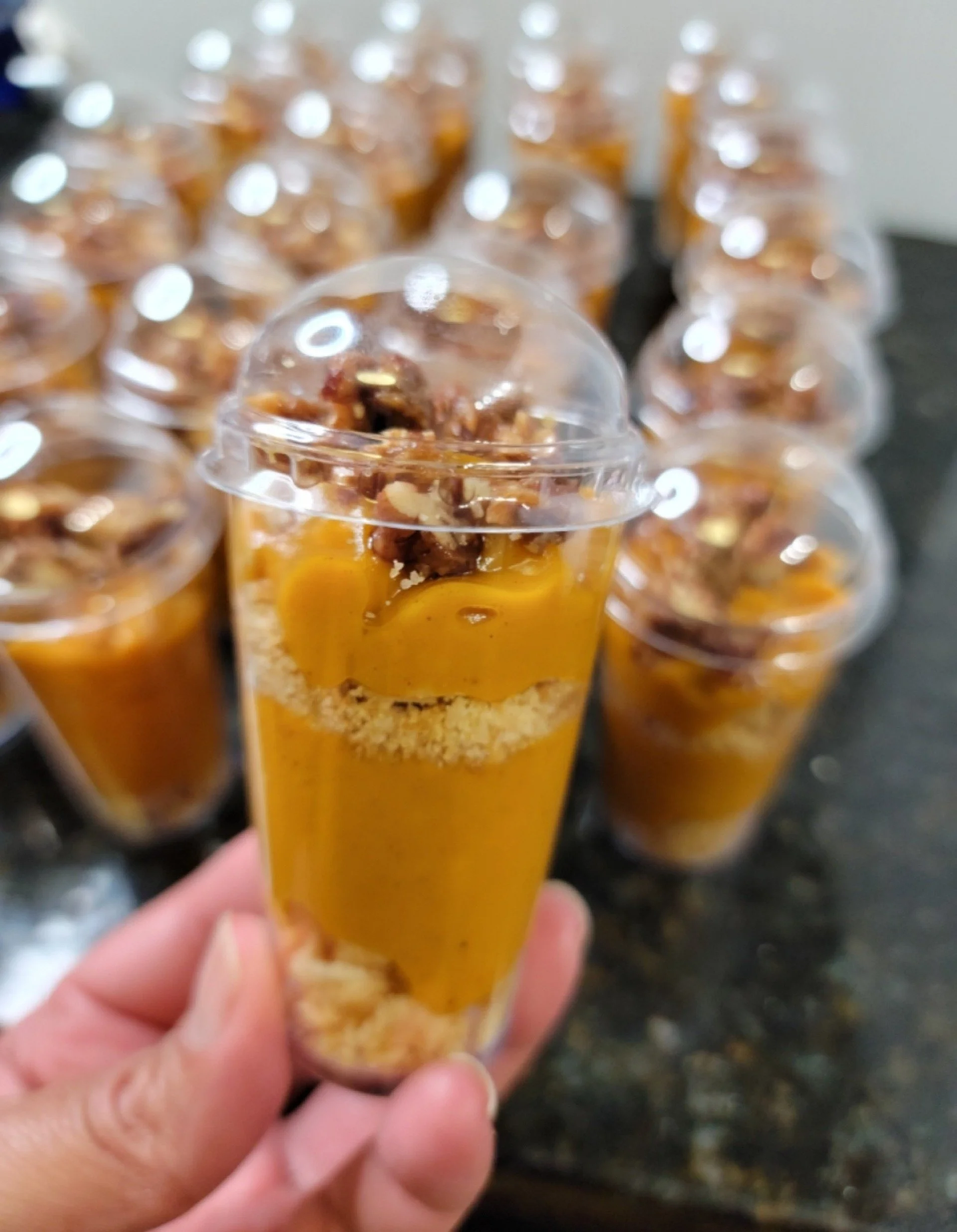 Hand holding a clear plastic cup filled with caramel-flavored dessert topped with chopped nuts, with several similar cups in the background.