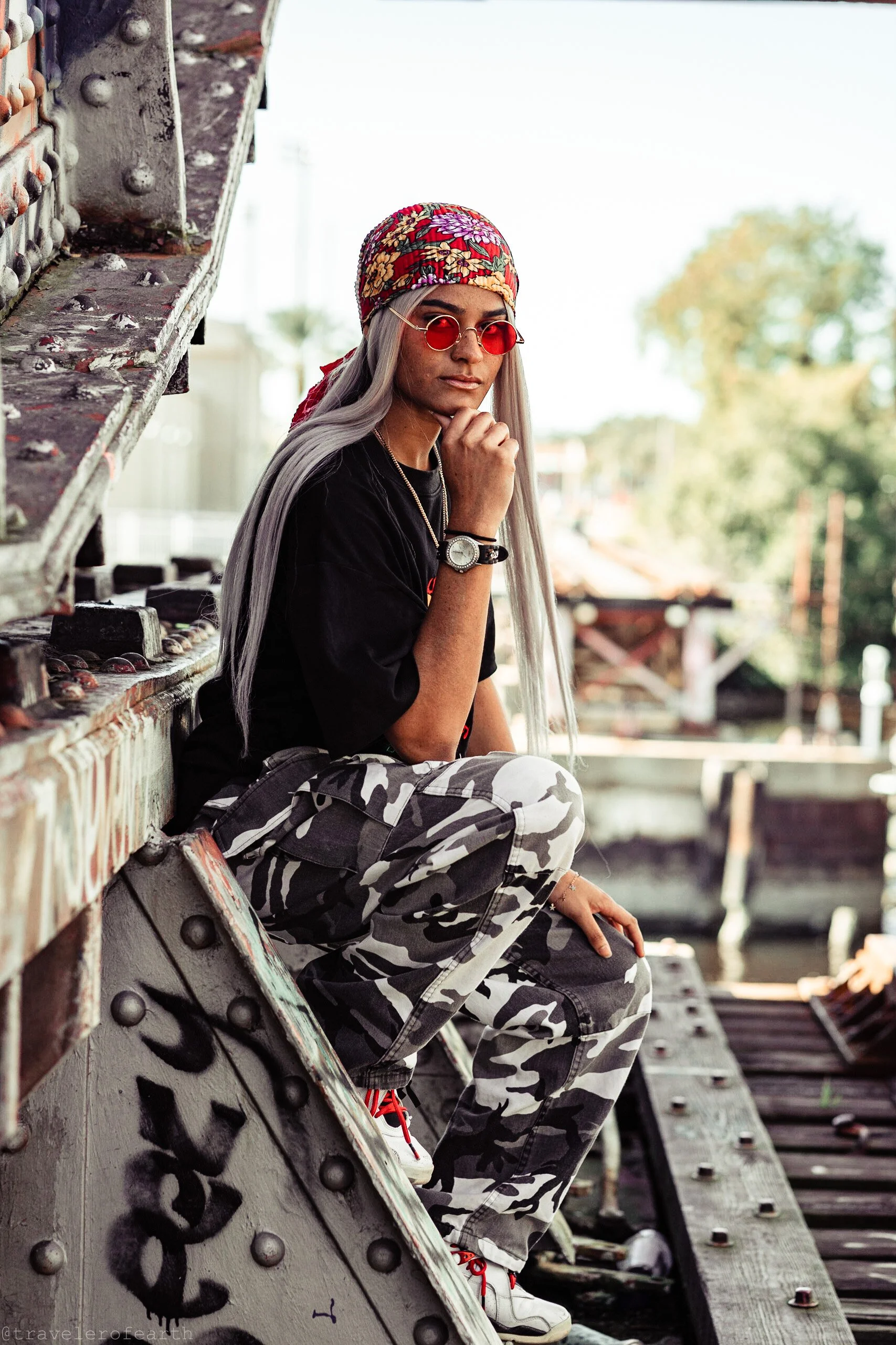 A woman with long silver hair, wearing a colorful bandana, red-tinted sunglasses, black t-shirt, gray camouflage cargo pants, and white sneakers. She is sitting on a graffiti-covered metal structure, resting her chin on her hand, with a watch and bra