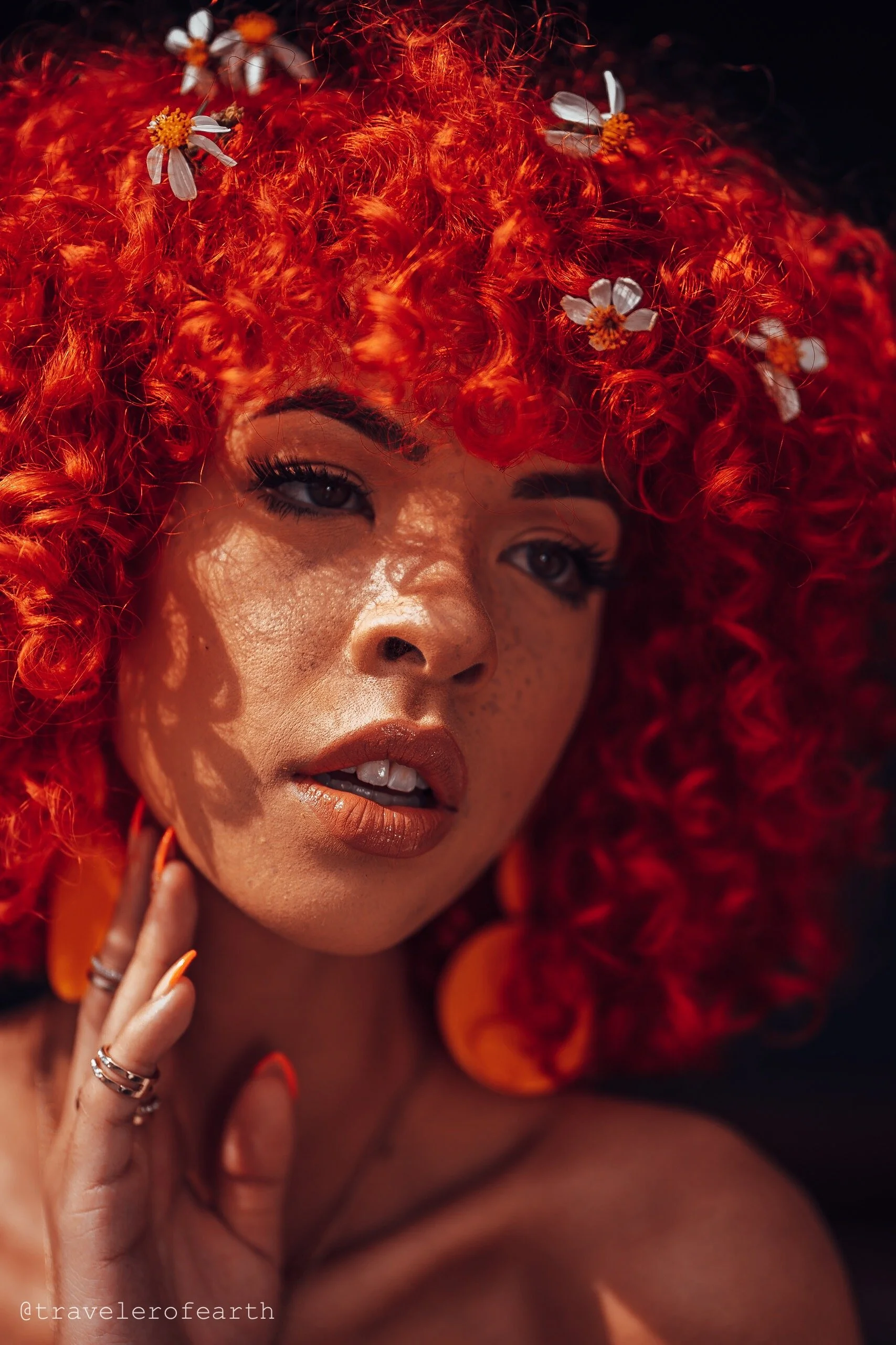 Close-up portrait of a woman with curly red hair adorned with small white flowers, wearing orange earrings, with natural makeup and freckles.