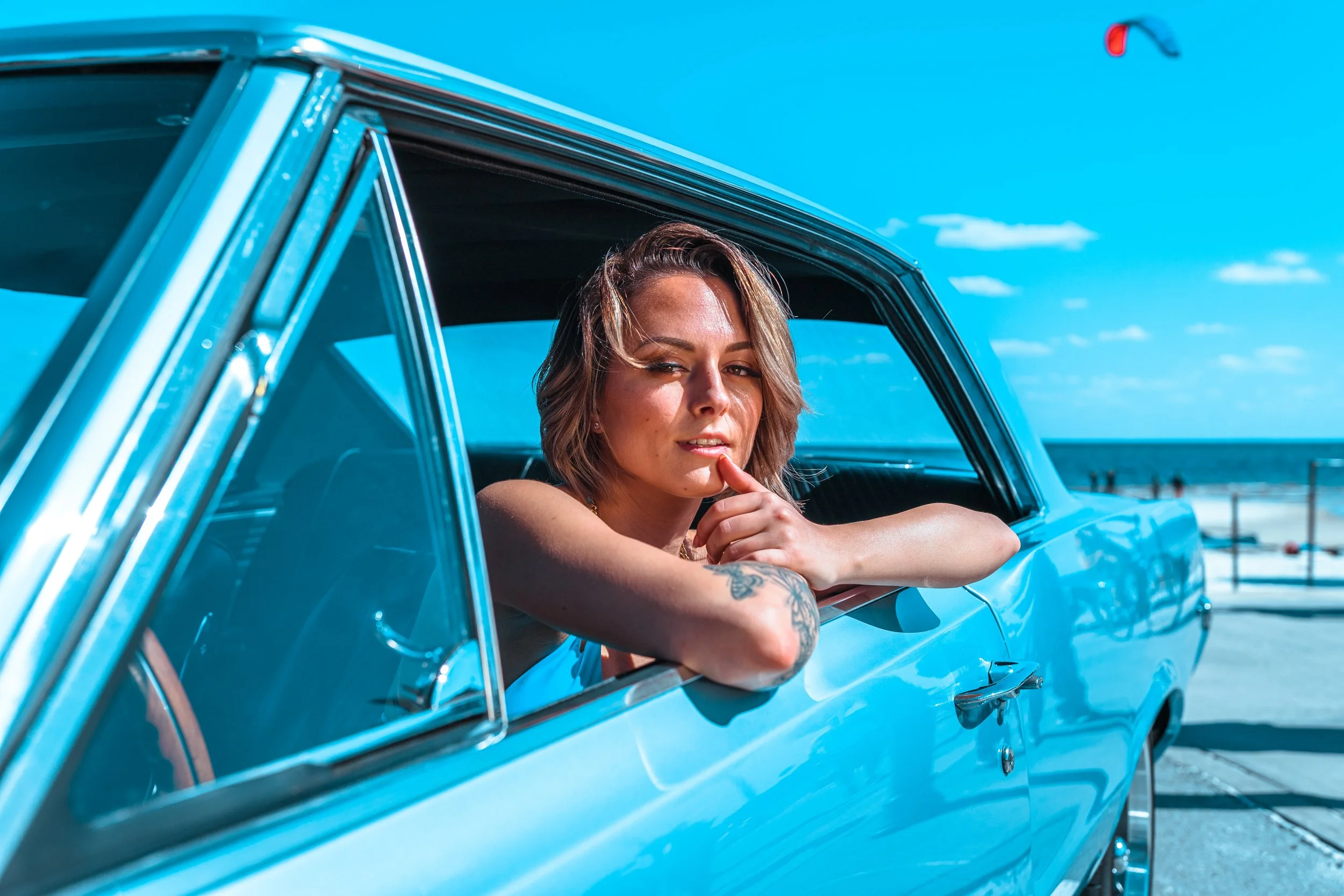 A woman with short brown hair and tattoos on her arm leaning out of a blue vintage car window at the beach, with ocean and blue sky in the background.