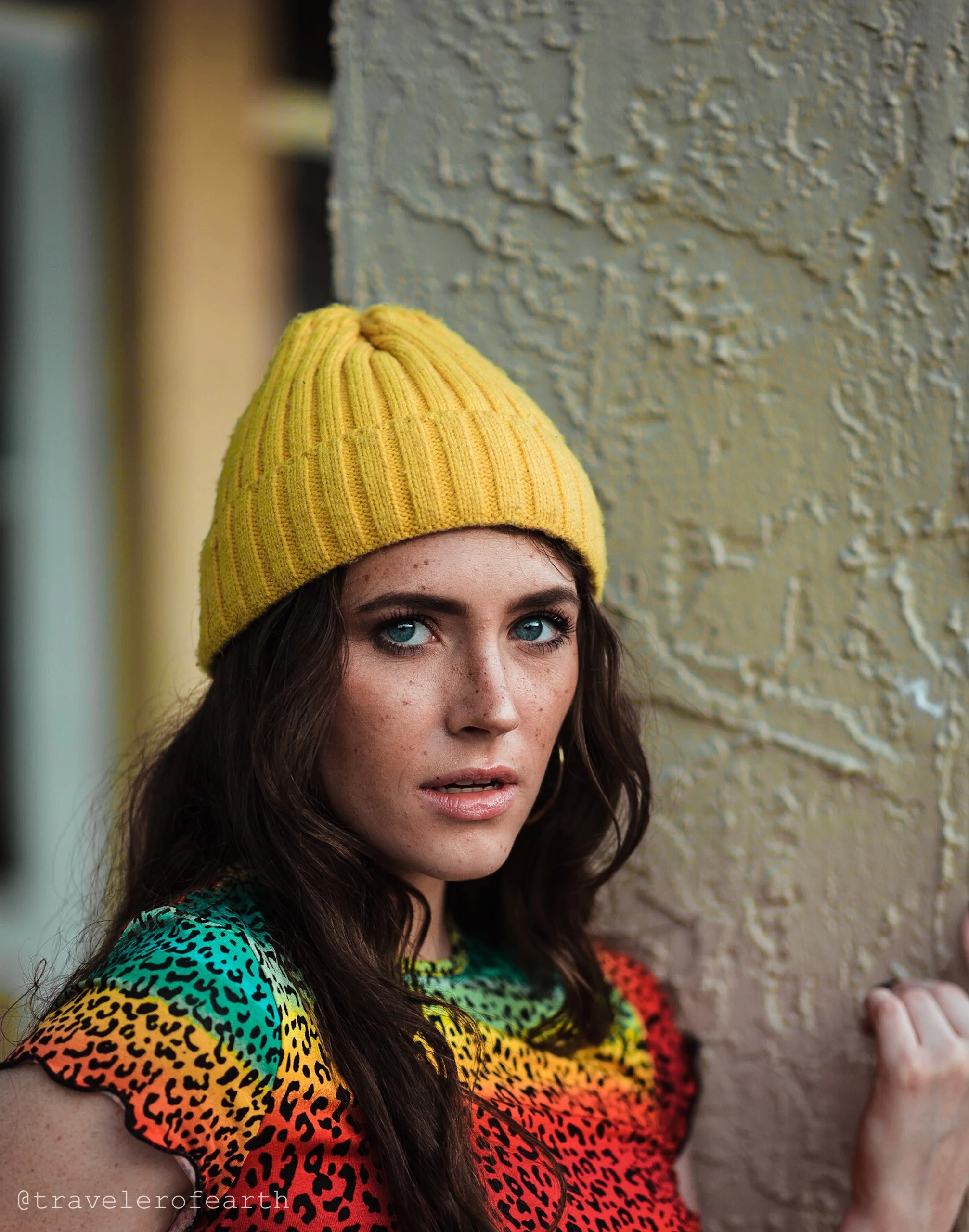 A young woman with blue eyes, freckles, and dark wavy hair wearing a yellow knit beanie and a colorful leopard print top, standing near a textured beige wall.