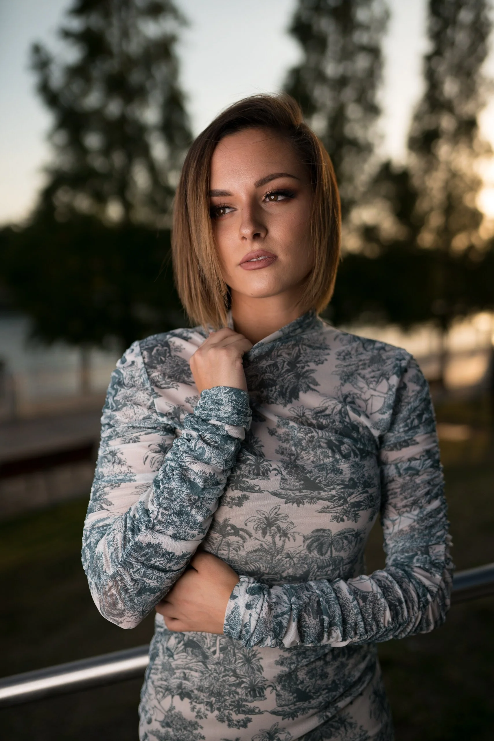 A woman with short brown hair and light makeup standing outdoors at sunset, wearing a patterned long-sleeve top with a tropical scene. She has her right hand on her neck and her left arm across her waist, looking contemplative.