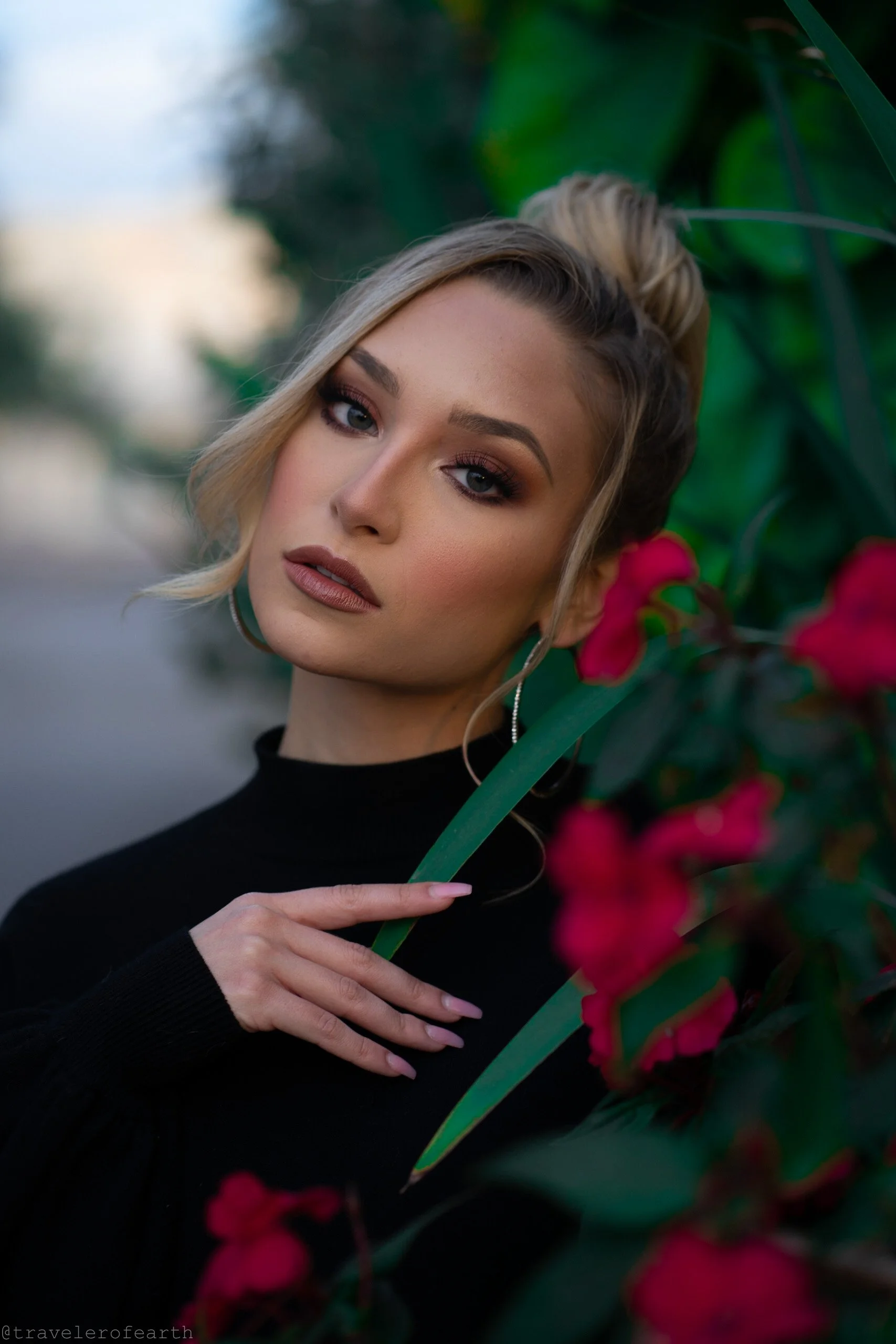 Portrait of a woman with blonde hair and makeup, wearing earrings, in a black top, with pink flowers and green leaves in the background.