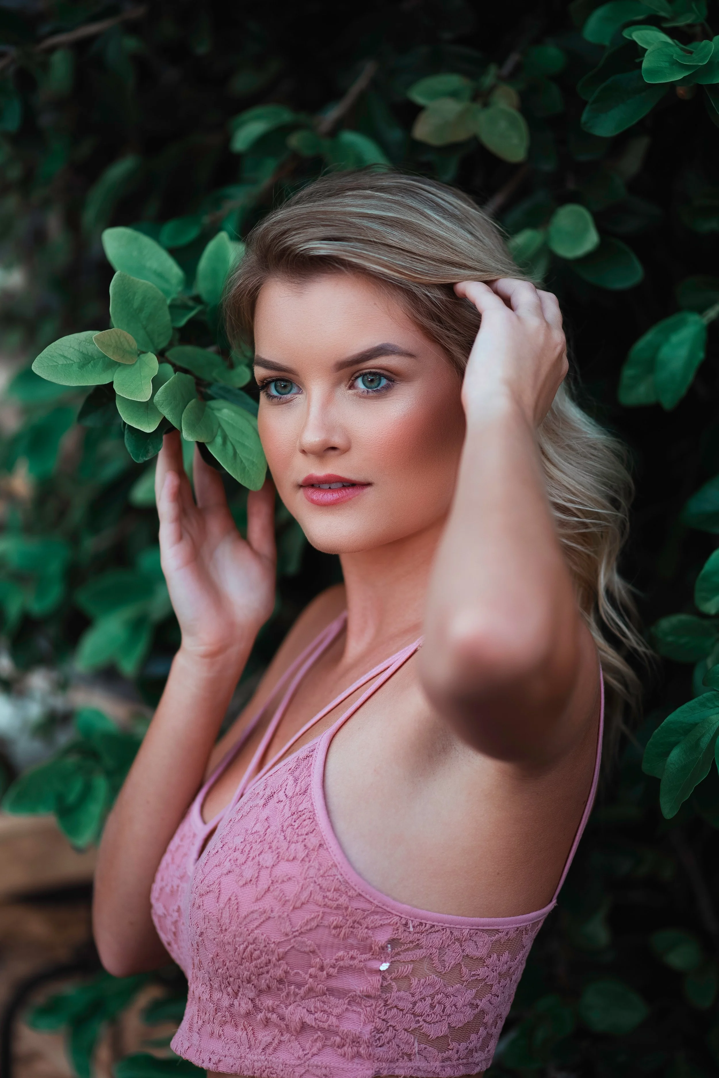A young woman with blonde hair and blue eyes wearing a pink lace top standing among green leaves.