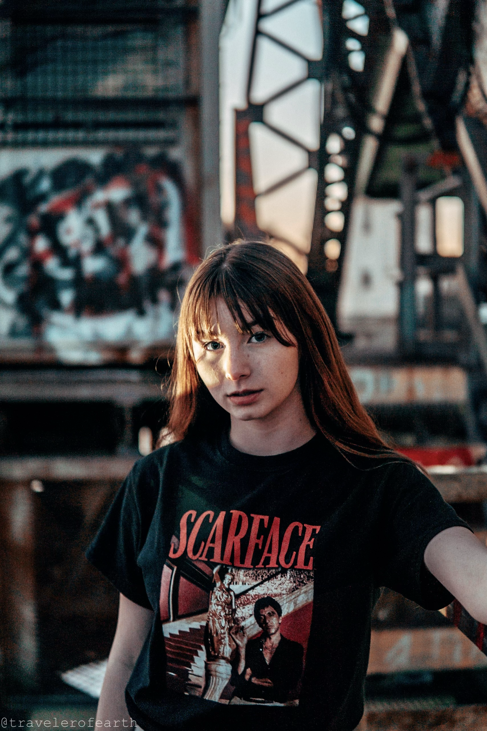 A young woman with long reddish-brown hair and bangs wearing a black 'Scareface' T-shirt with a graphic design, standing in an industrial setting with graffiti and metal structures in the background.