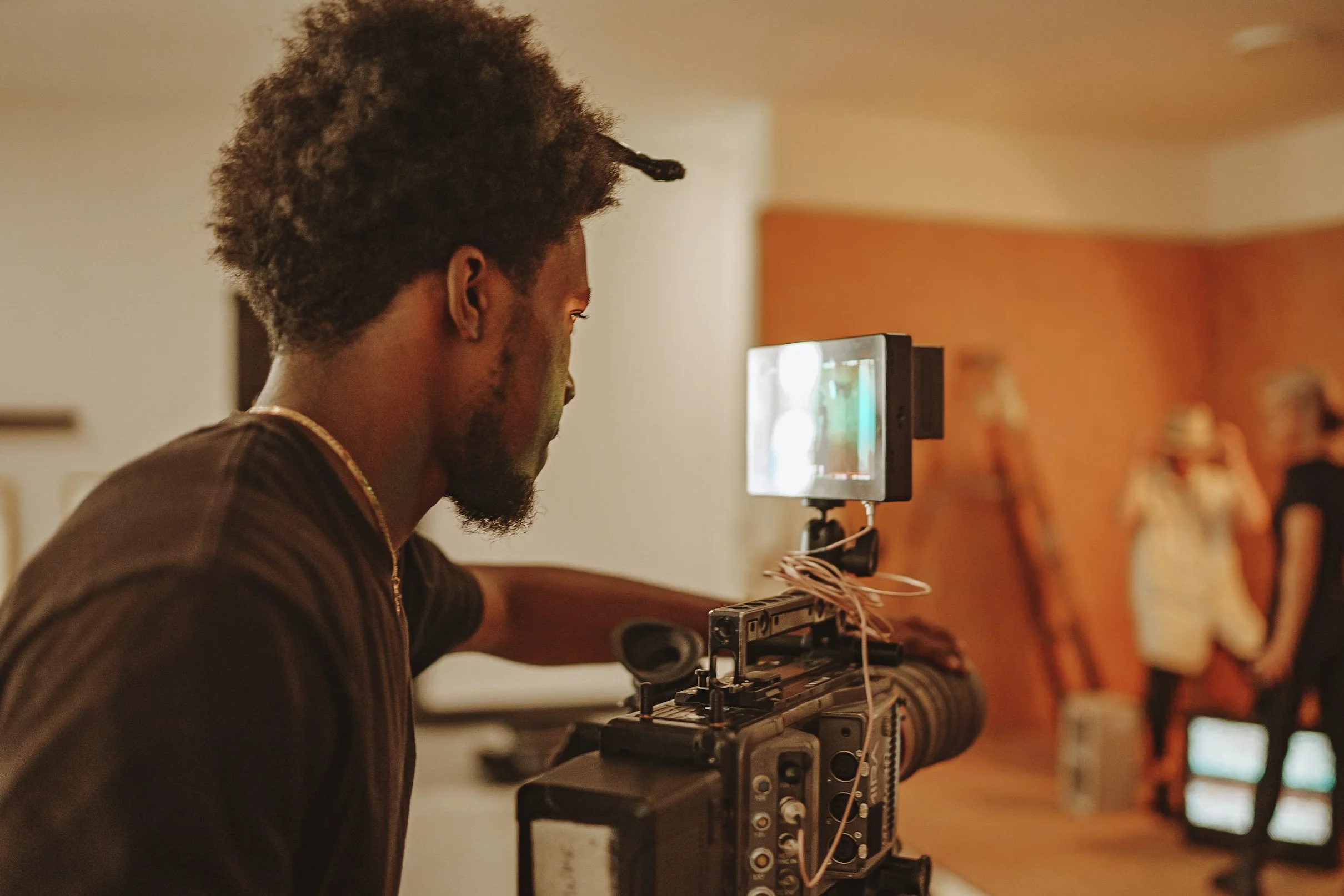 A man working with a professional video camera, monitoring the shot on a small screen, in a room where two women are holding hands and talking in the background.