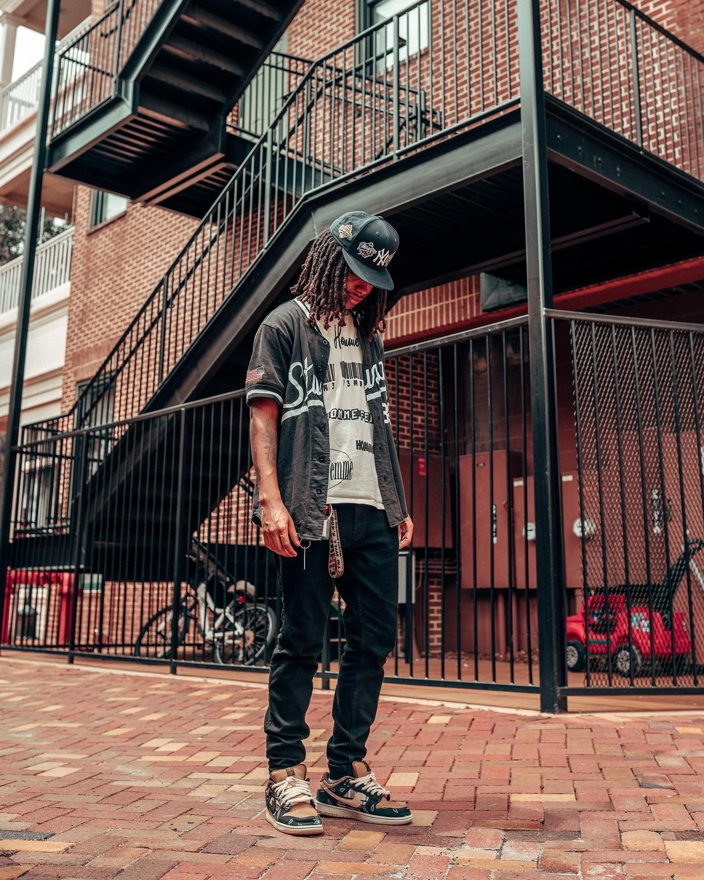 A man with dreadlocks wearing a baseball cap, black and grey jacket, dark jeans, and sneakers standing on a brick sidewalk in front of a black metal staircase and brick building.