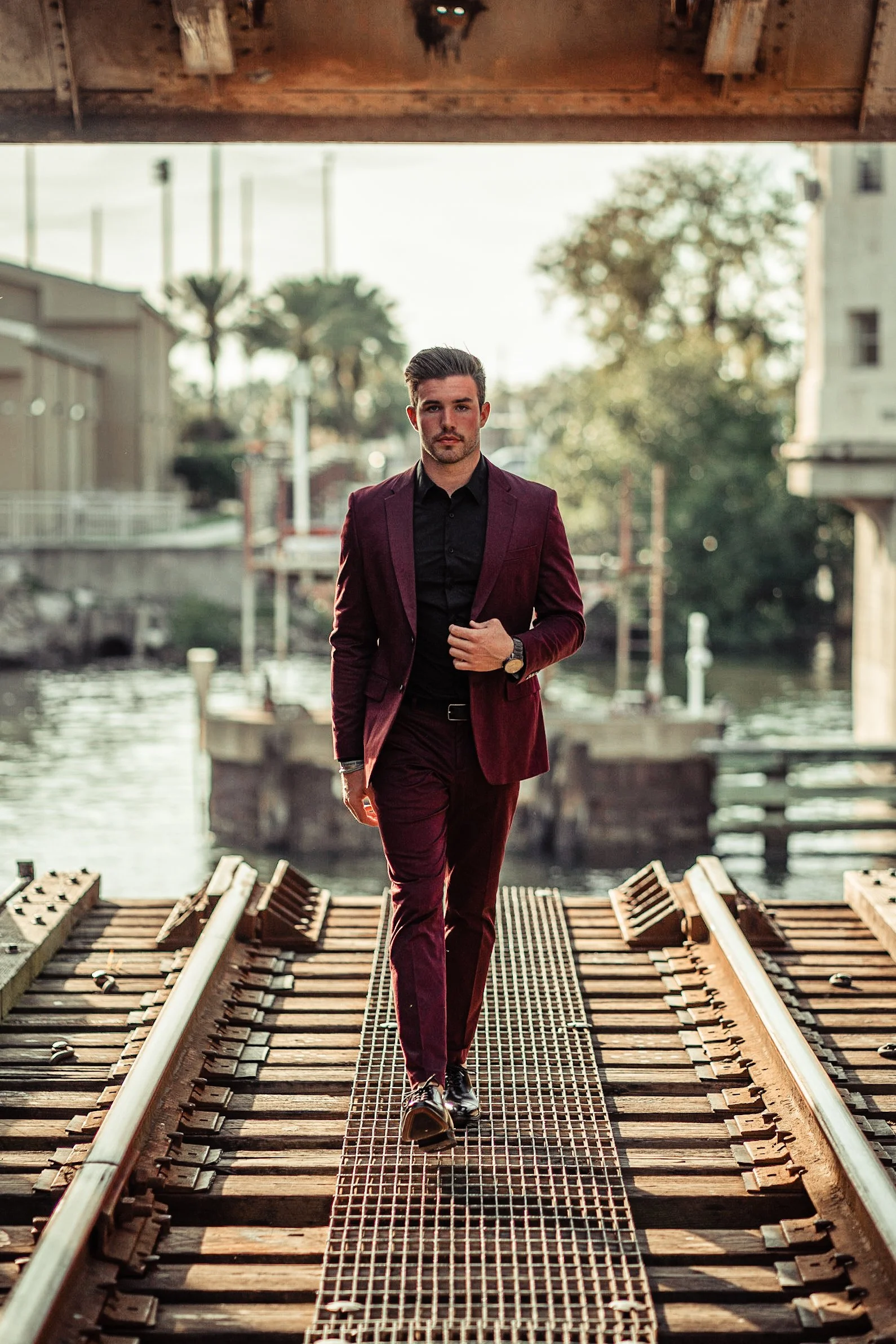 A man in a burgundy suit and black shirt walking on a railroad bridge over water outdoors.
