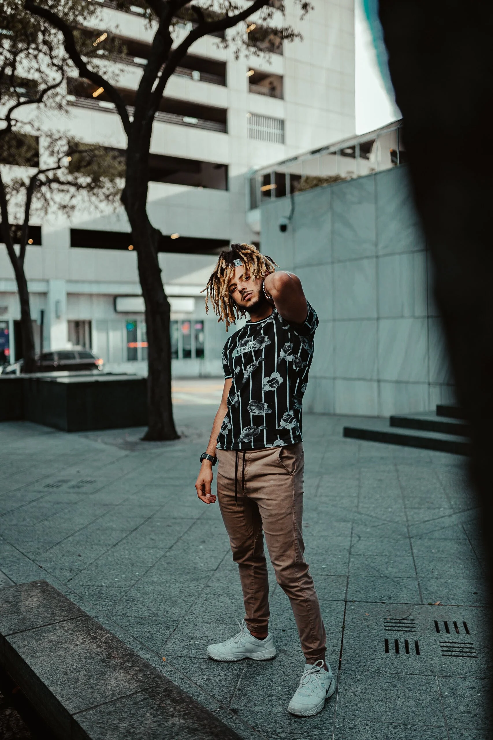 A young man with blonde dreadlocks standing outdoors on a city sidewalk, wearing a black t-shirt with a floral pattern, beige joggers, and white sneakers. He has a watch on his left wrist and is looking at the camera with one hand behind his head, ag