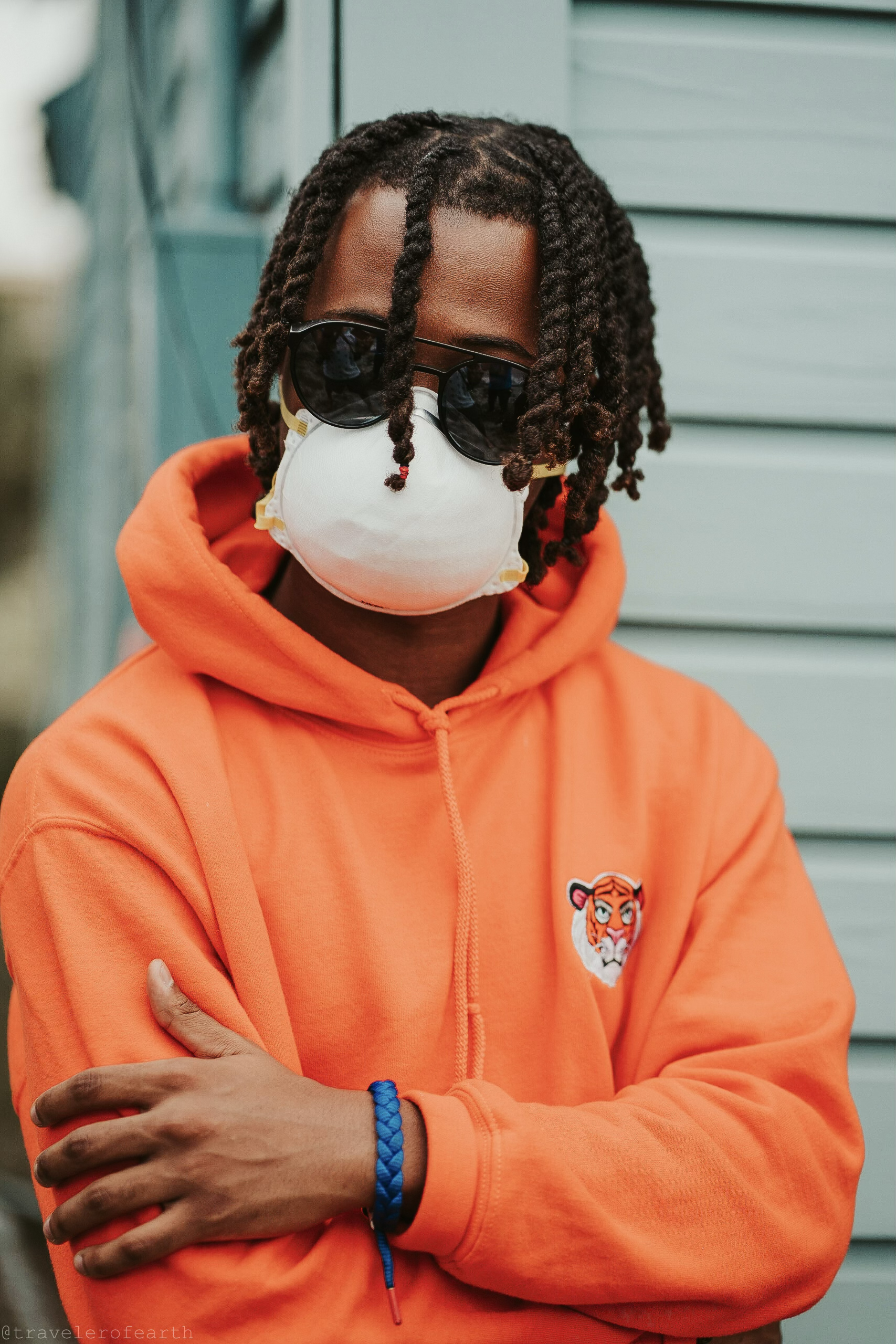 Person with dreadlocks wearing sunglasses, a face mask, an orange hoodie with a tiger logo, and a blue bracelet standing with arms crossed.