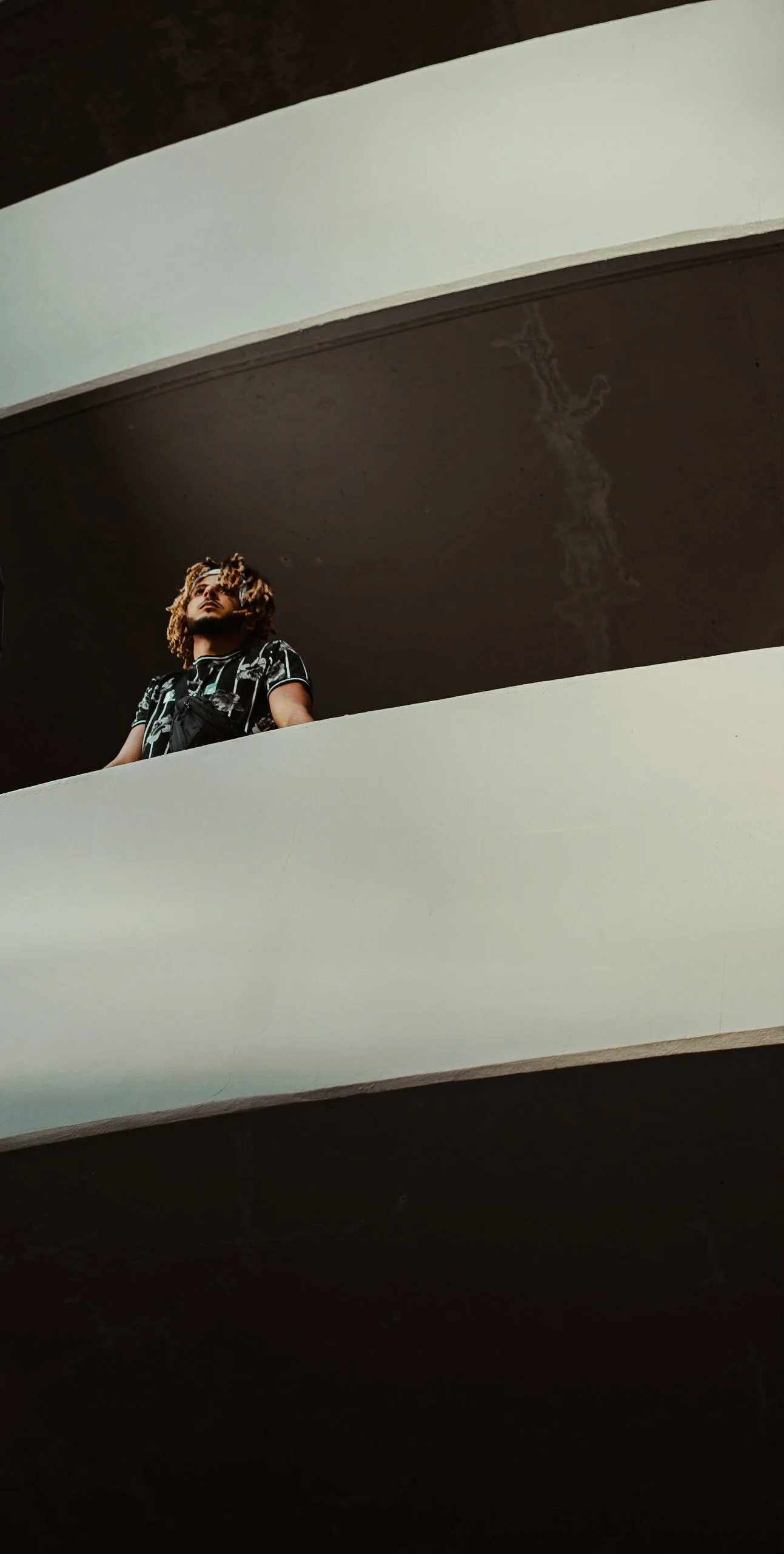 Man with dreadlocks standing on a balcony looking up, seen from below.