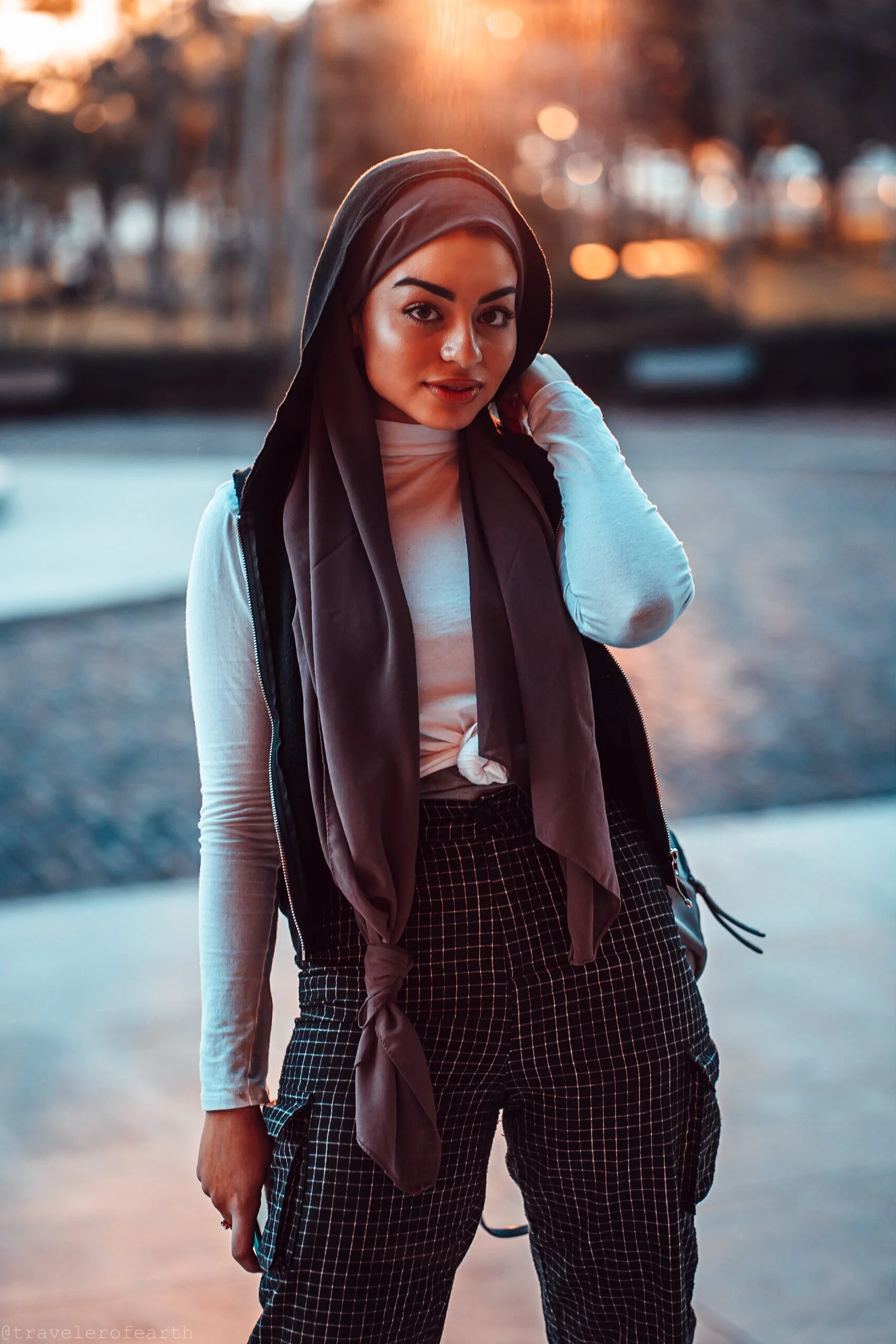 A young woman with dark hair and eyebrows, wearing a black headscarf, white long-sleeve shirt, black checkered cargo pants, and a black vest, standing outdoors during sunset.