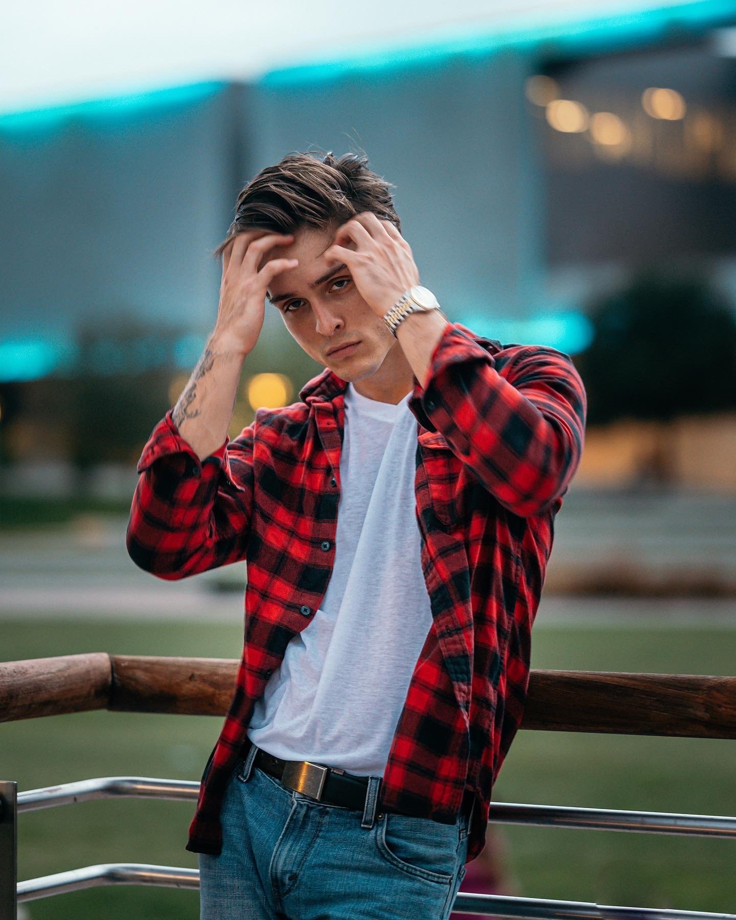 Young man with styled hair, wearing a red plaid shirt over a white T-shirt, and a wristwatch, standing outdoors with a metal railing behind him, appearing frustrated or deep in thought.