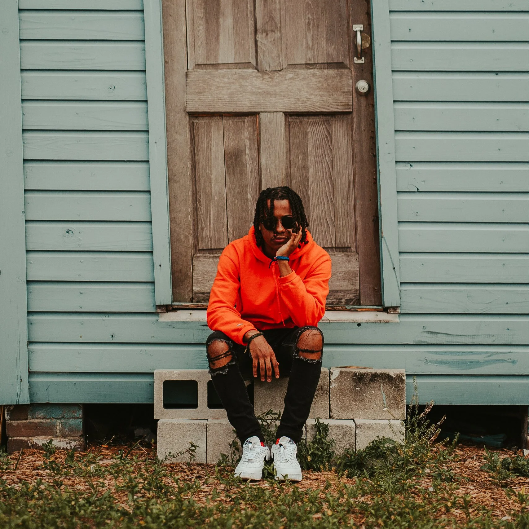 A person wearing an orange hoodie, ripped black jeans, and white sneakers sitting on cinder blocks in front of a weathered wooden door with blue siding on a house.