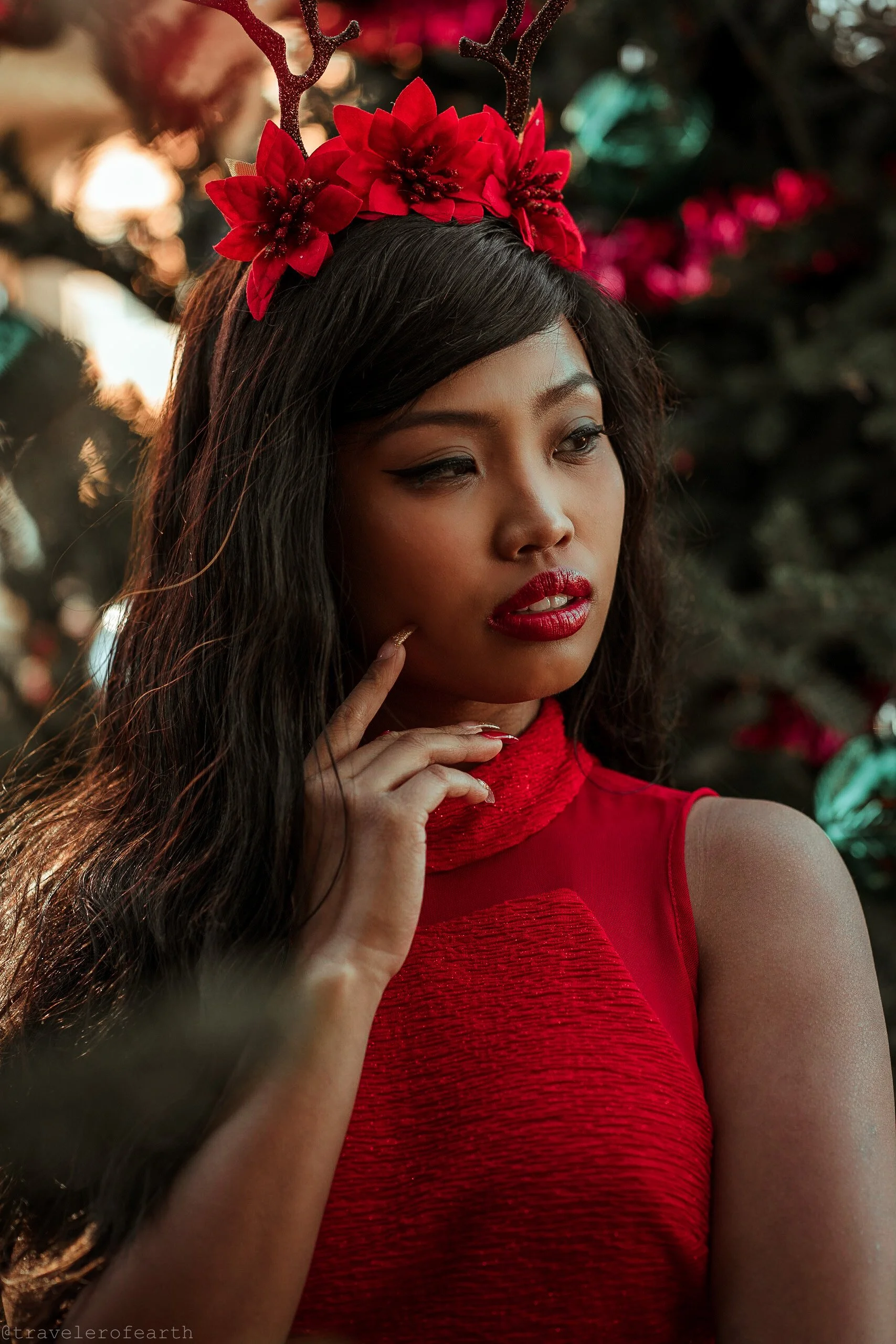 A woman wearing festive reindeer antlers with red poinsettia flowers, red lipstick, and a red sleeveless dress, standing outdoors near Christmas trees decorated with ornaments and lights.
