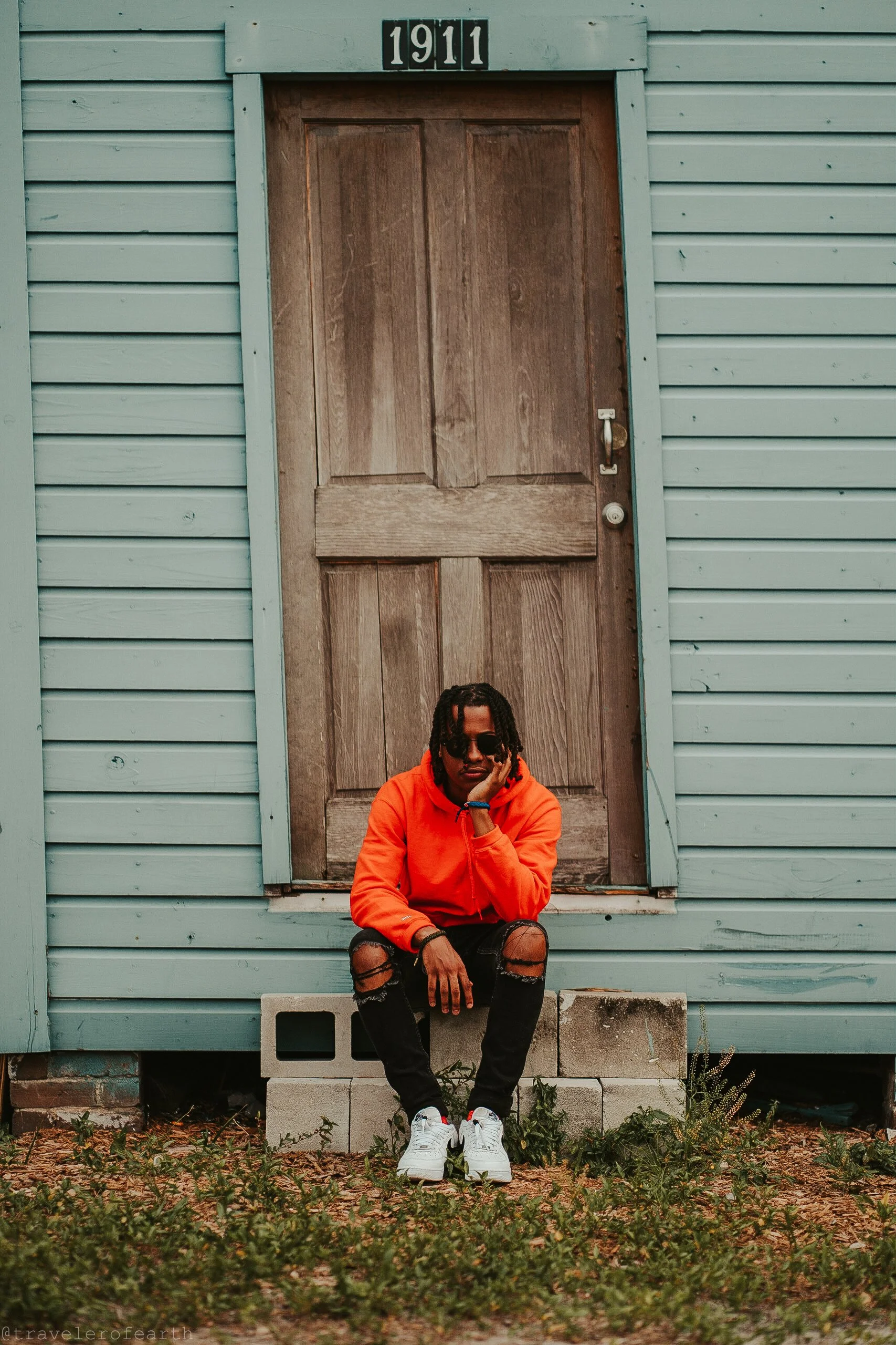 A person wearing an orange hoodie, ripped black jeans, and white sneakers sitting on concrete blocks in front of a weathered wooden door on a light blue house, with the number 1911 above the door.
