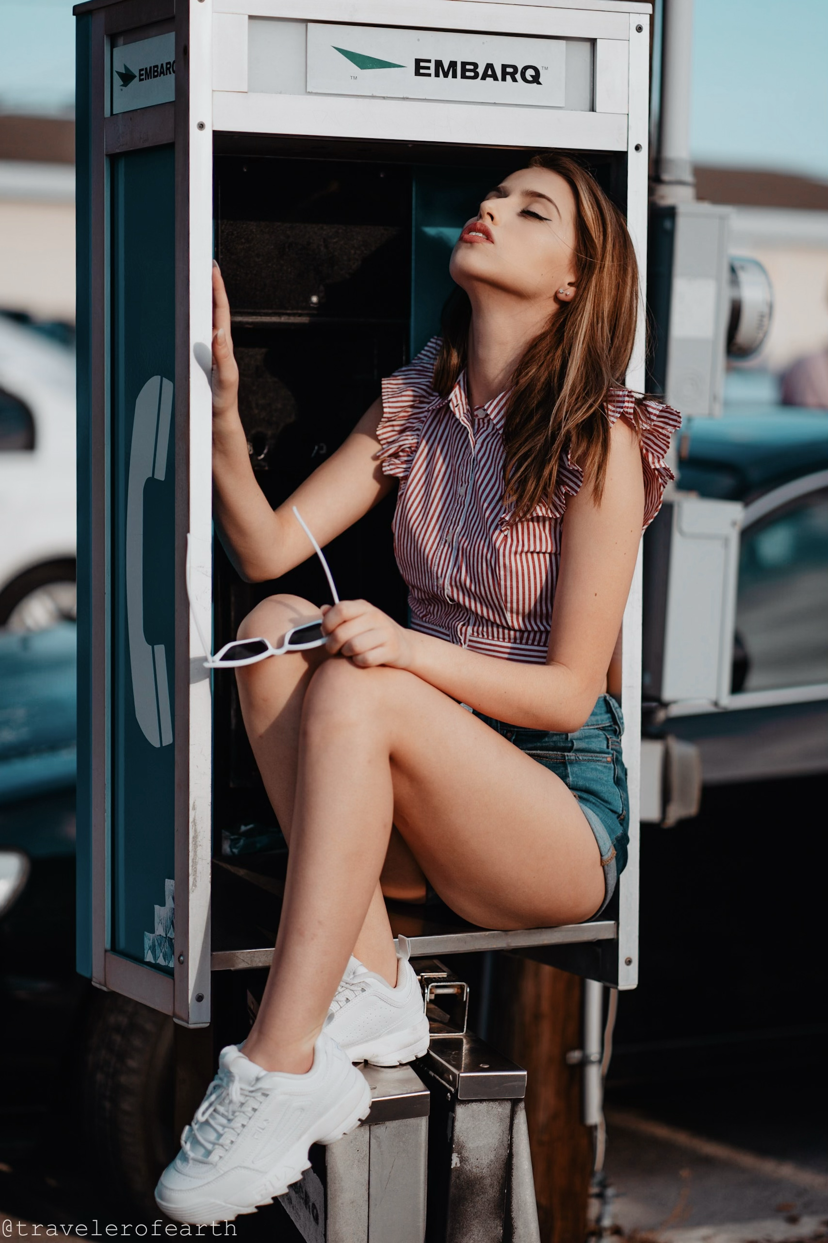 A young woman with brown hair sits inside a vintage payphone booth, holding a pair of white sunglasses, with her eyes closed and head tilted back in a relaxed pose during daylight hours.