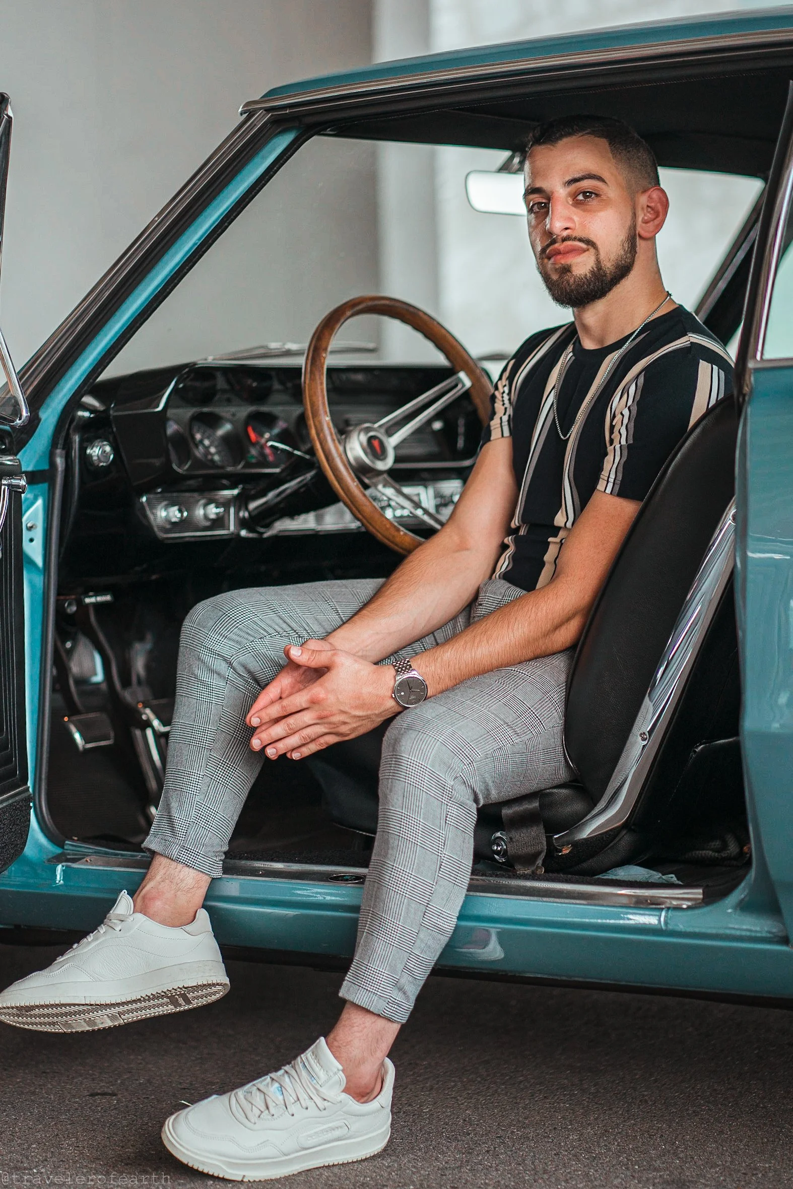 A young man sitting in the driver's seat of a vintage blue car, with one leg outside the car, wearing white sneakers, plaid pants, a striped shirt, and a watch, inside a well-lit indoor space.