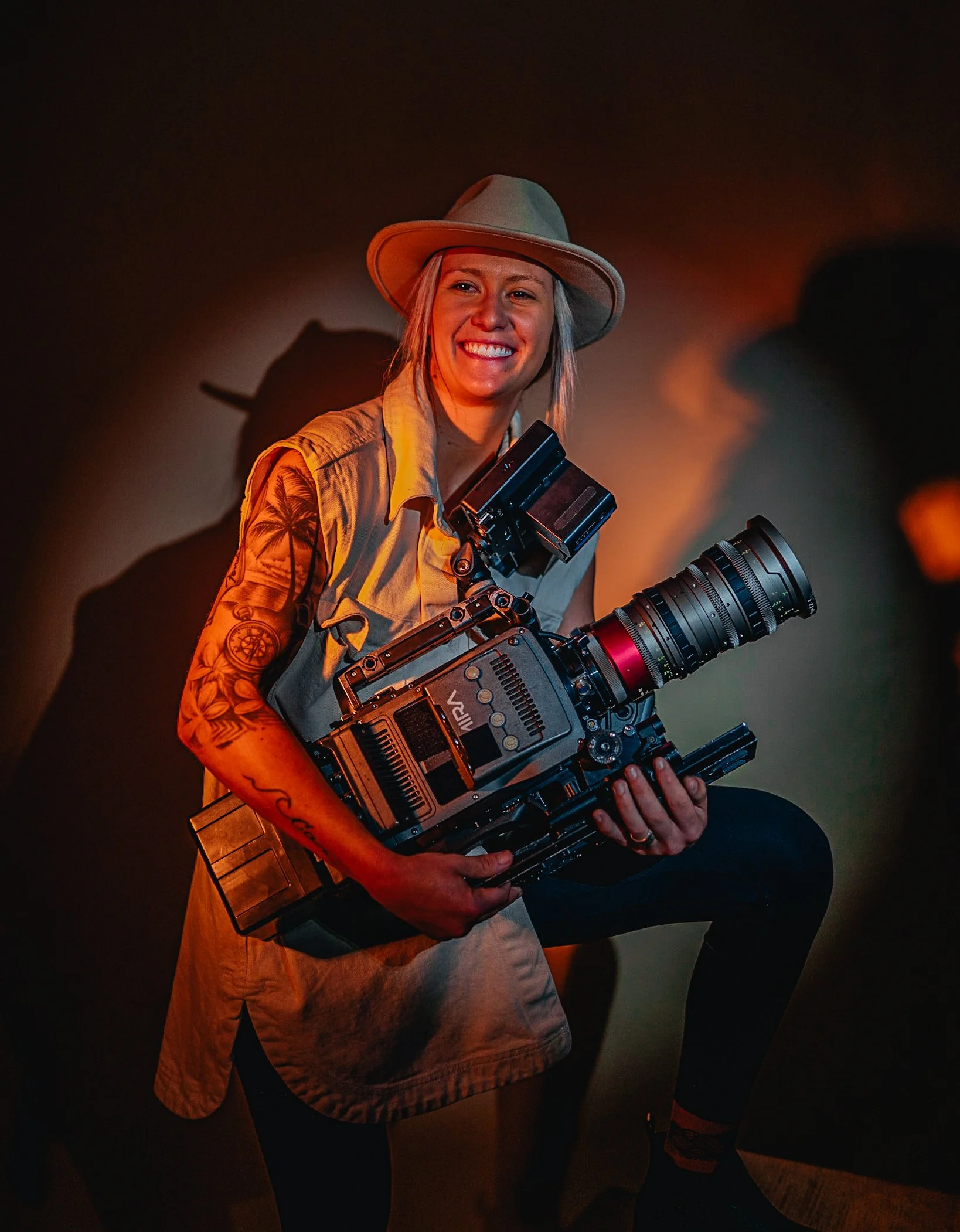 Woman in a beige wide-brim hat holding a large professional film camera, smiling, wearing a tan shirt and dark pants, with tattoos on arms, in a dimly lit environment.