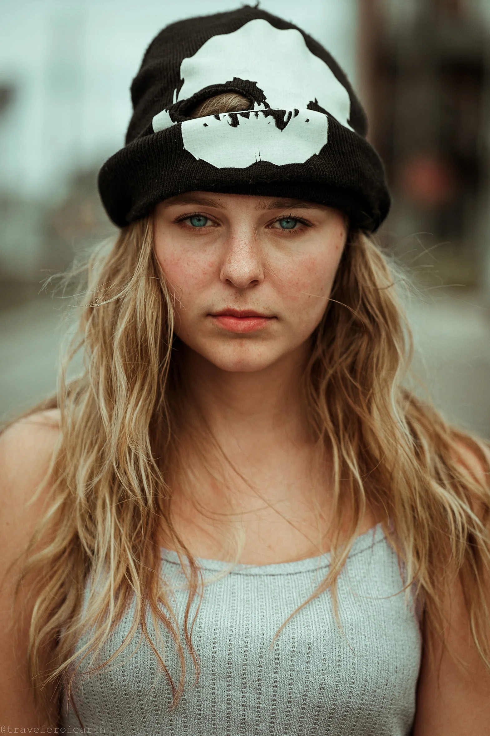 A young woman with long blond wavy hair and blue eyes wearing a black beanie hat with a white skull design, looking directly at the camera.