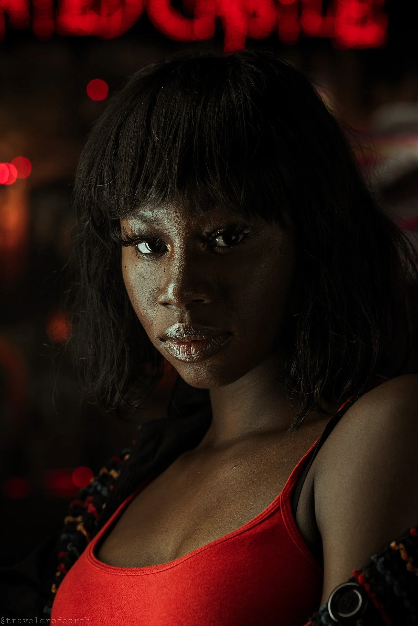 A portrait of a woman with dark skin and shoulder-length black hair, wearing a red top, in a dimly lit setting with red lights in the background.