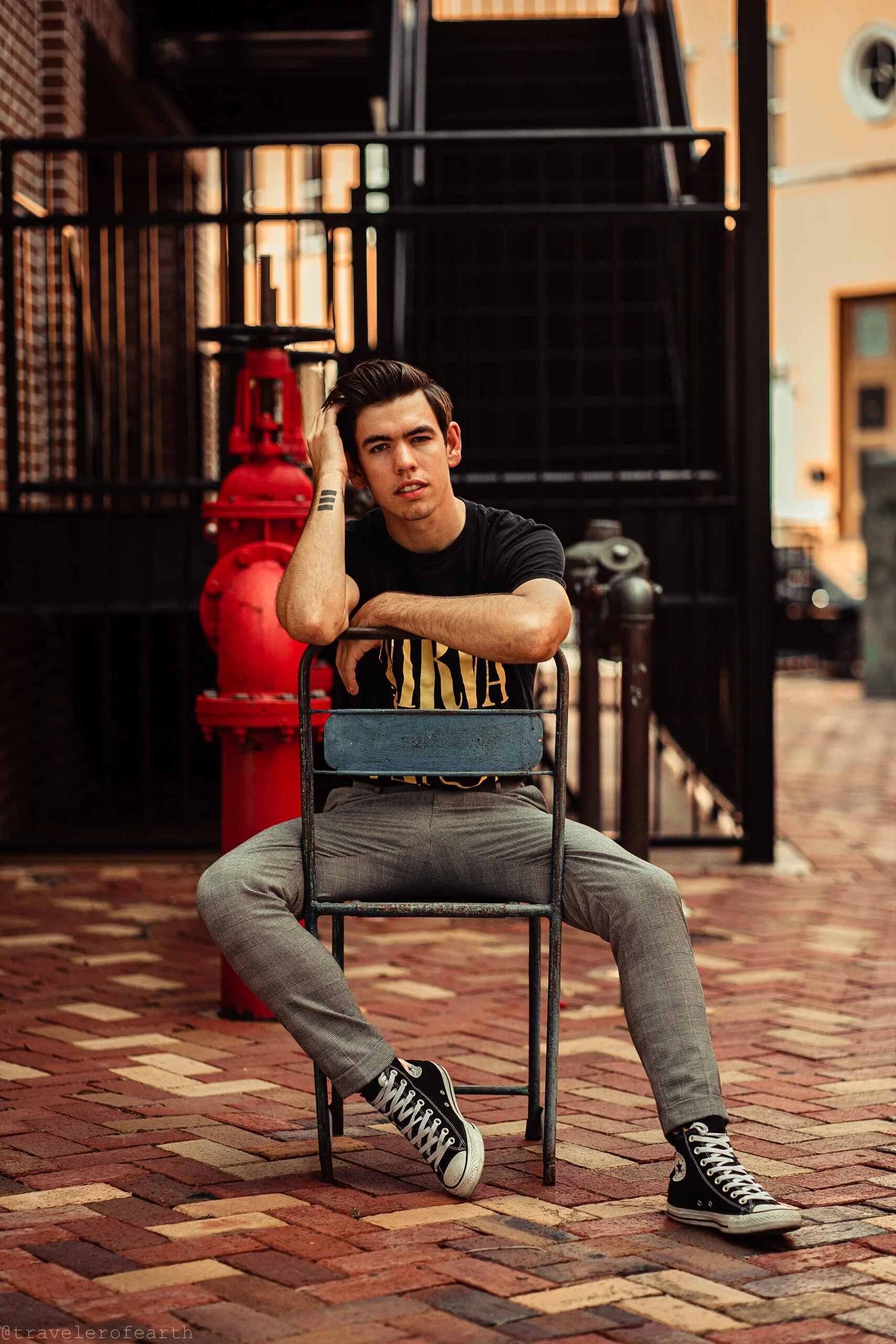 A young man in a black T-shirt, gray pants, and black high-top sneakers sitting on a metal chair on a brick sidewalk, with his arm resting on the backrest and one hand touching his hair, looking at the camera. Behind him are a red fire hydrant and bl