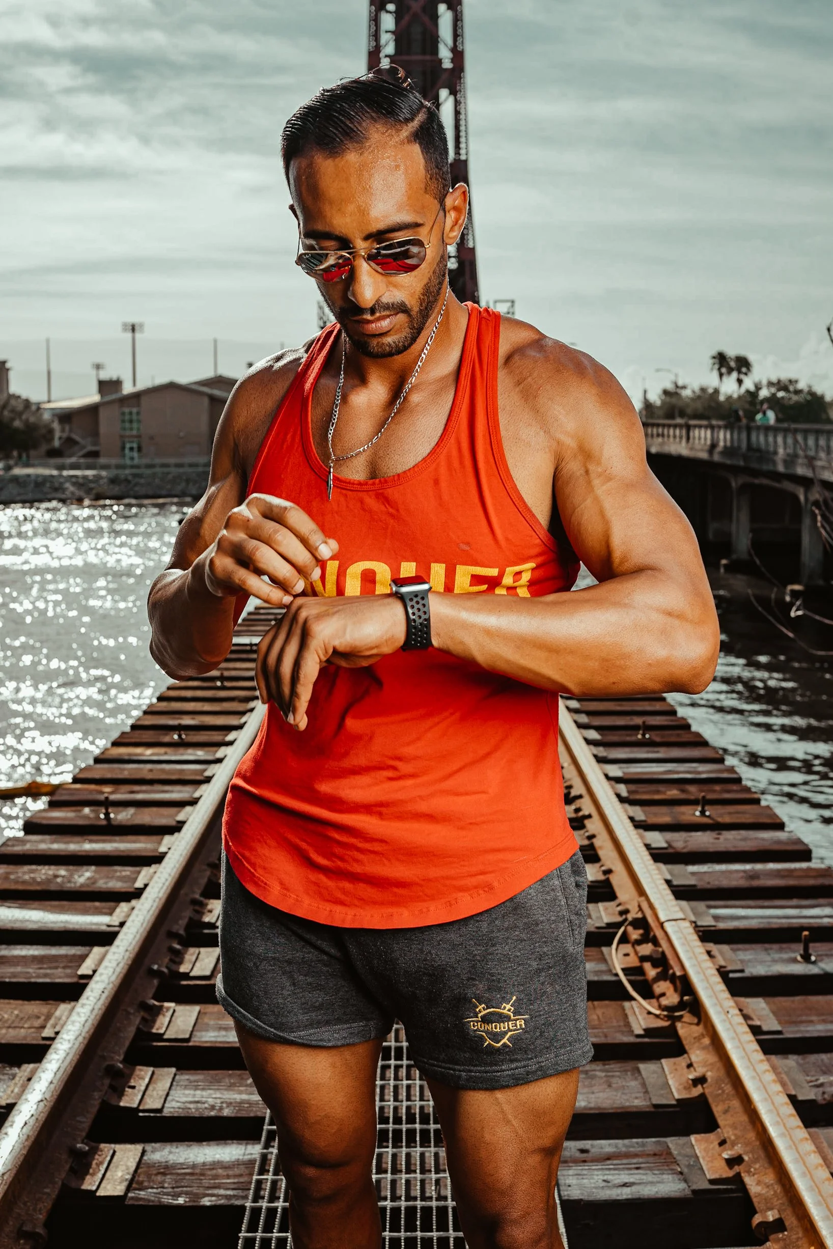 A man in sportswear is standing on railroad tracks near a body of water, looking at his smartwatch while checking the time or activity, with the Golden Gate Bridge in the background.
