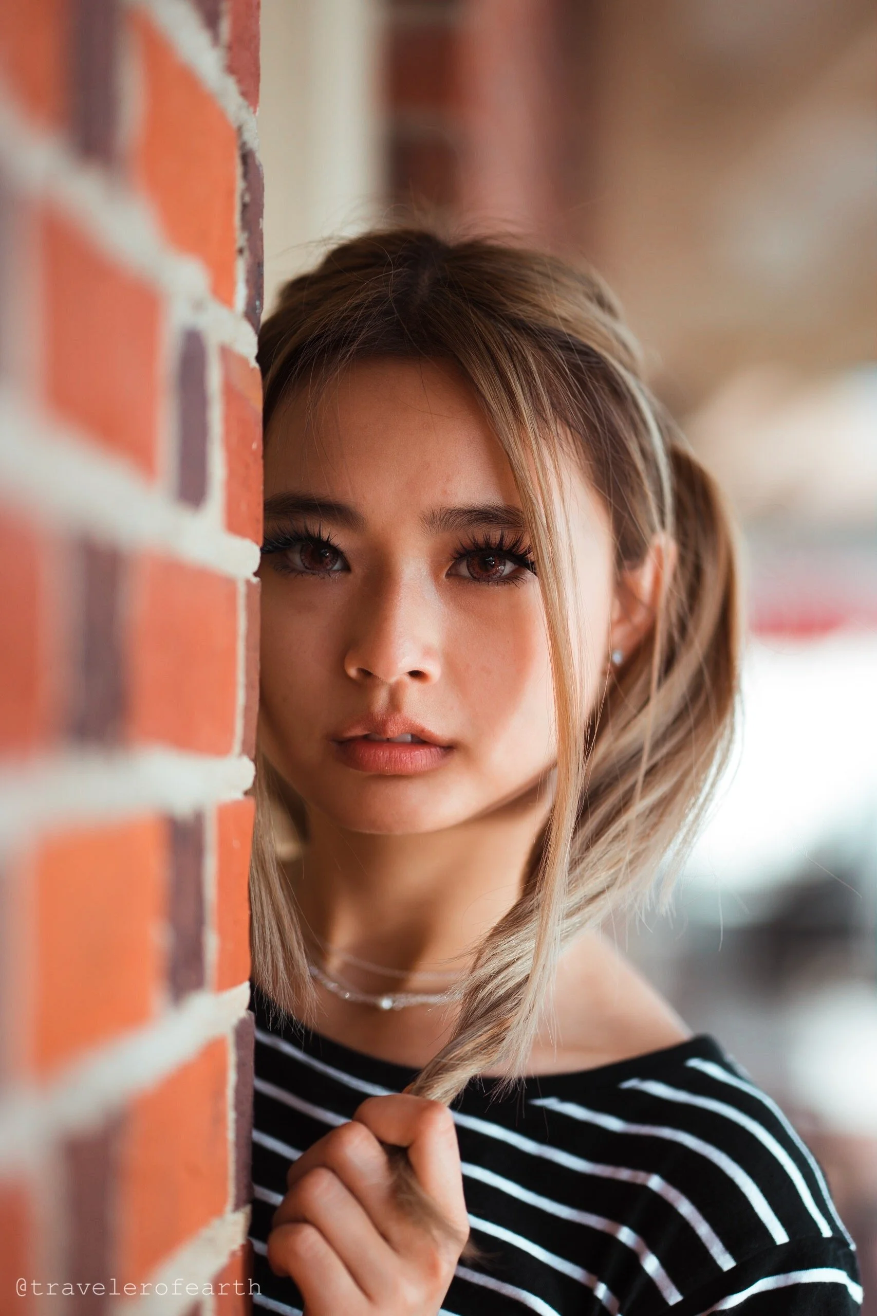A young woman looks directly at the camera, standing partly behind a brick wall, wearing a black and white striped shirt and a necklace.
