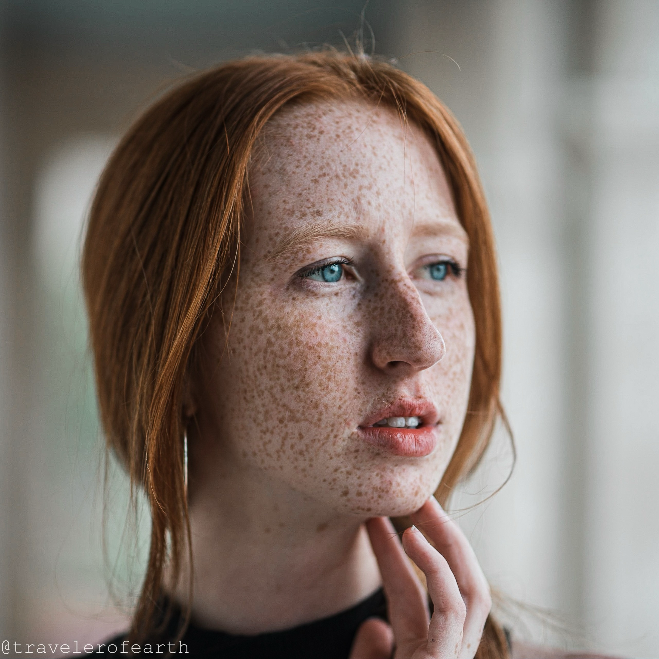 Close-up of a young woman with red hair and blue eyes, looking thoughtfully to the side with her hand near her chin.
