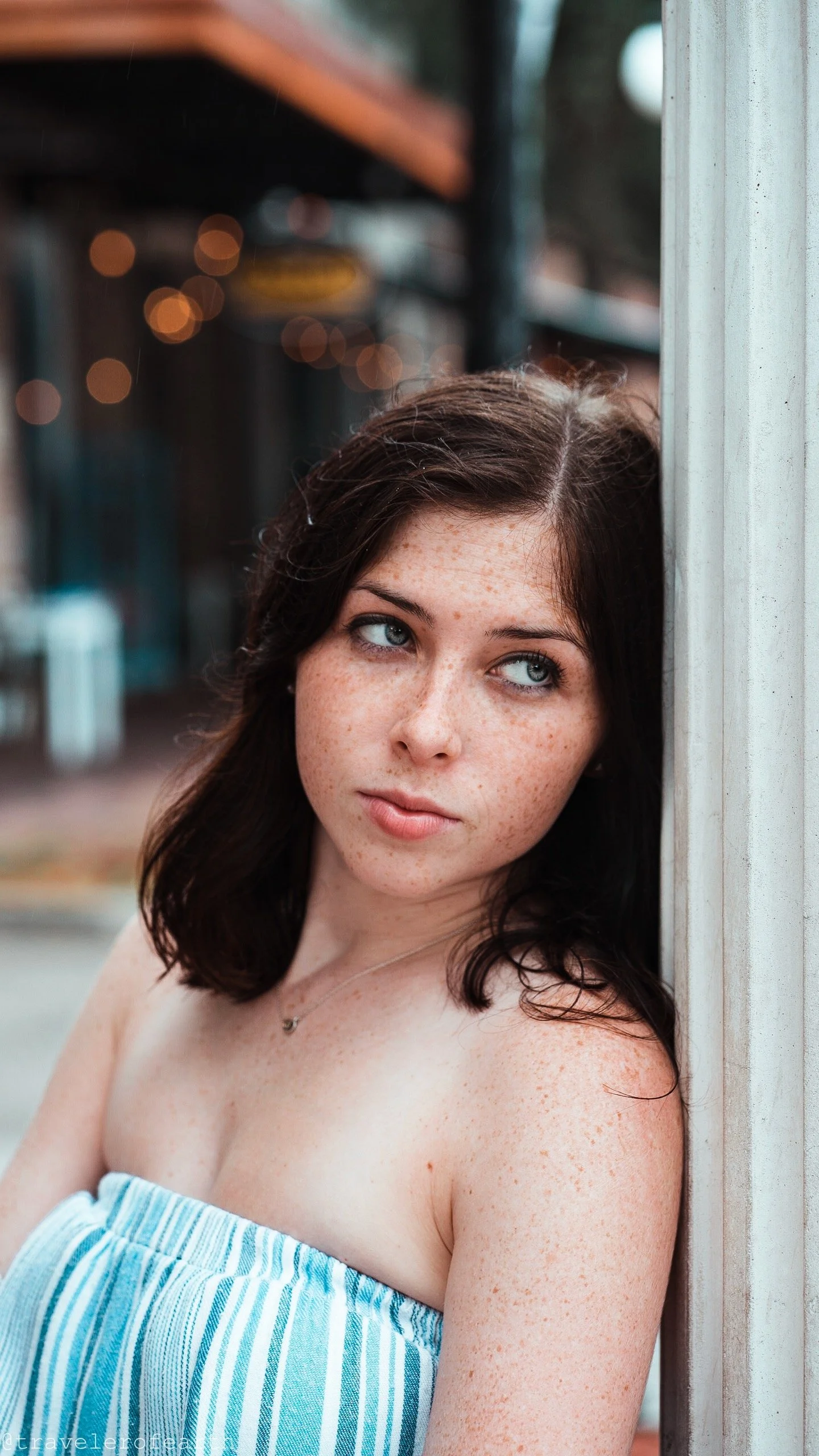 A young woman with dark brown hair, blue eyes, and freckles looking to her left while leaning against a wall.