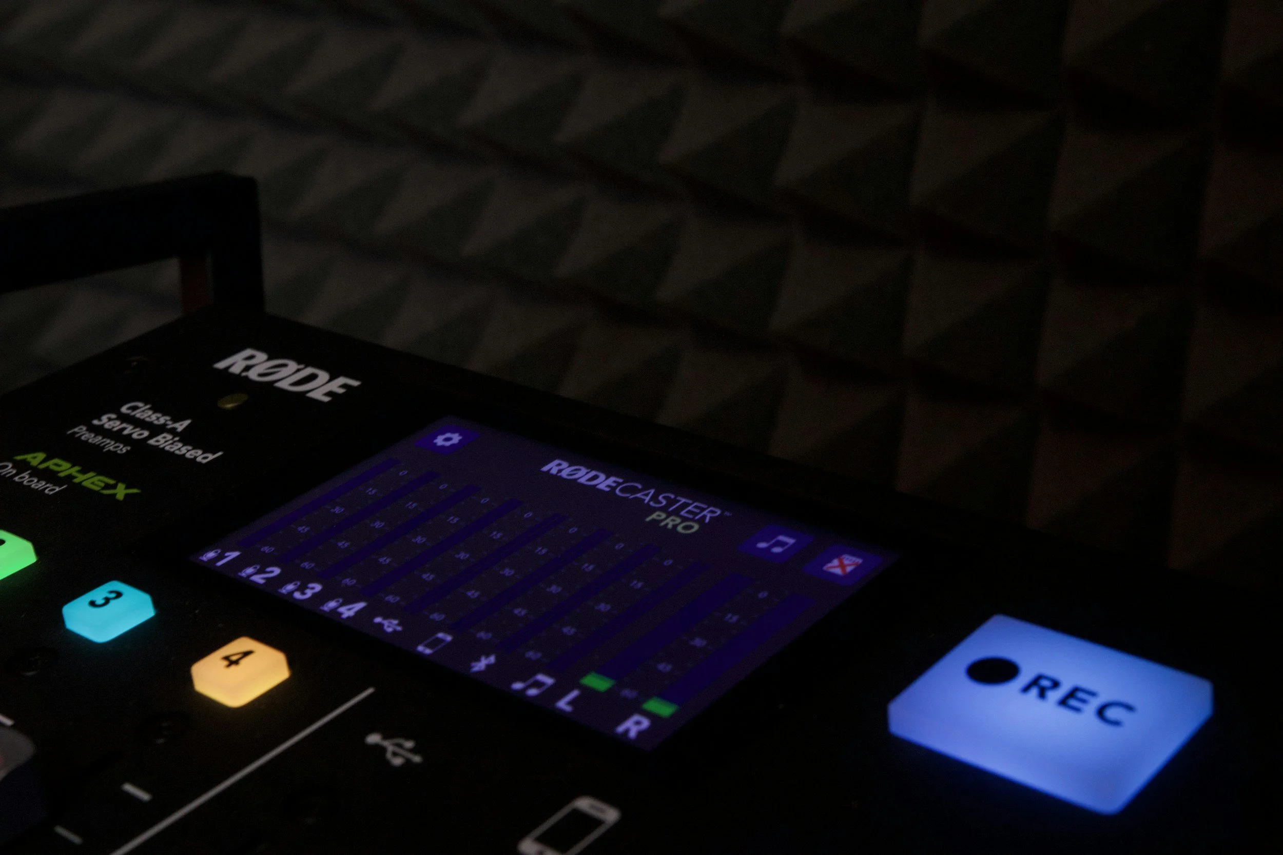 A close-up photo of a digital device screen displaying a software interface, with buttons lit in various colors on the left side, and the branding 'RODE' visible at the top. The background is dark with a patterned fabric surface.