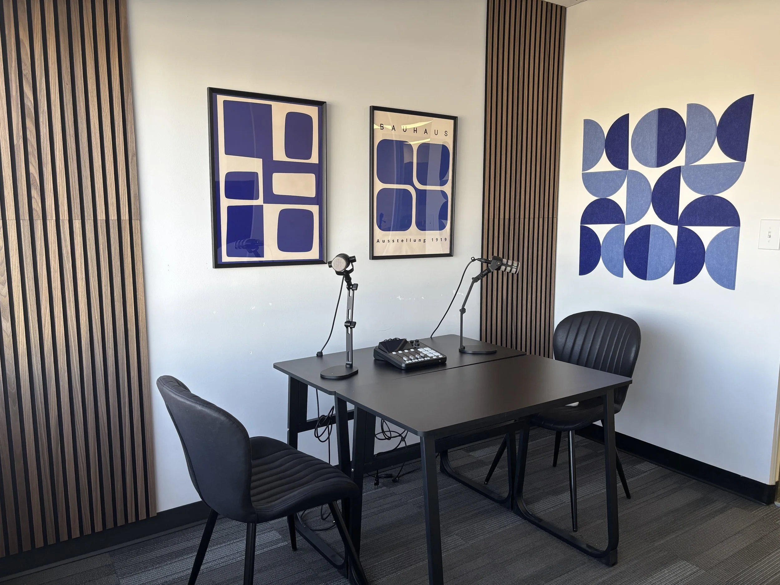 Professional podcast recording studio in Calgary with a black table, two microphones on desk stands, audio mixer, black chairs, acoustic wood slat wall panels, and modern blue abstract artwork.