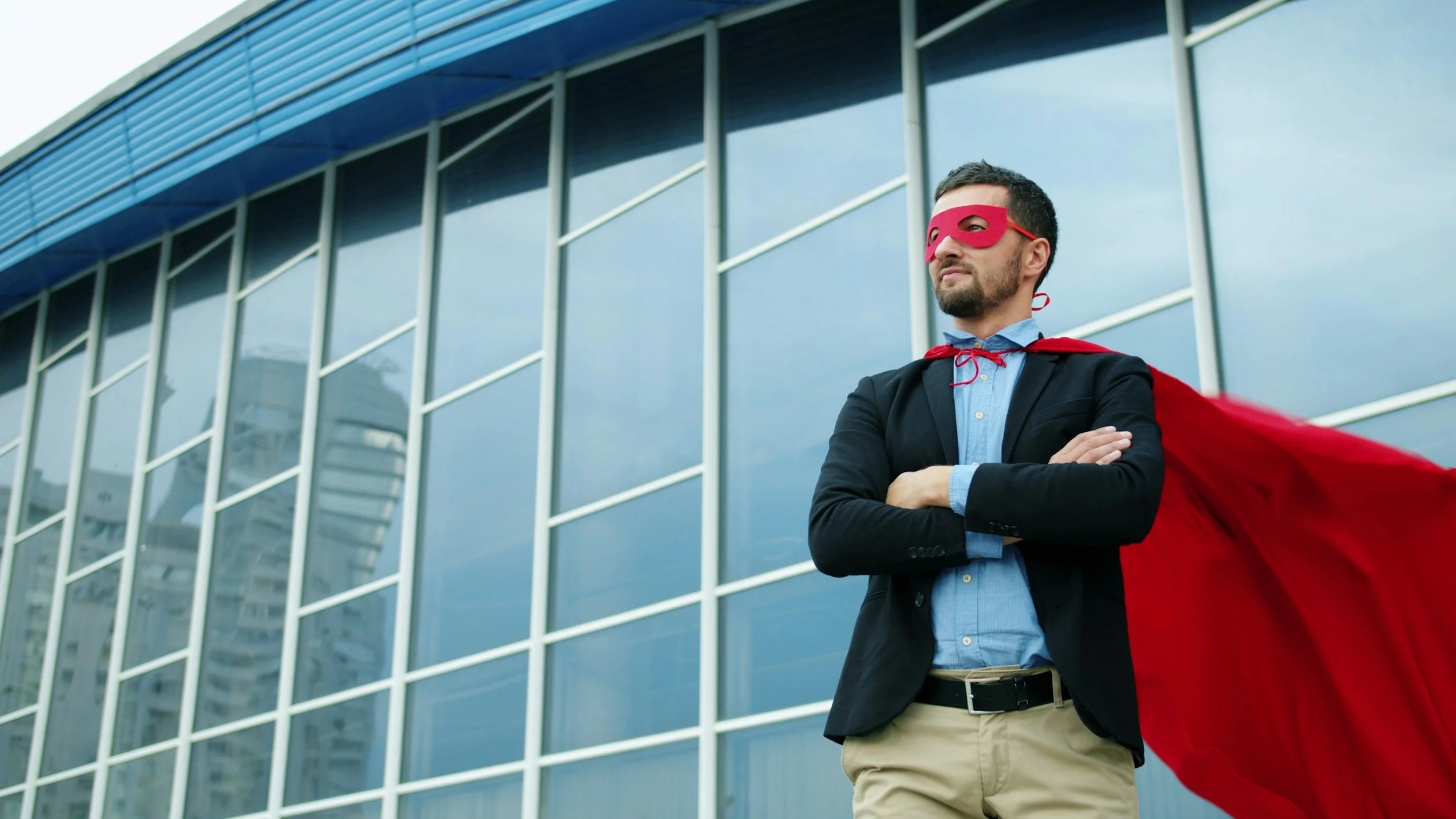 A man wearing a red superhero mask and red cape stands confidently with crossed arms in front of a modern glass building.