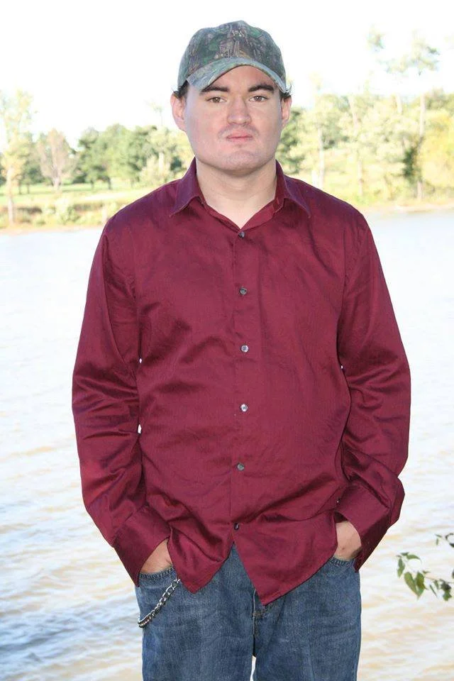 A young man with fair skin and dark hair wearing a camo baseball cap, a burgundy button-up shirt, and blue jeans, standing outdoors near a body of water with trees in the background.