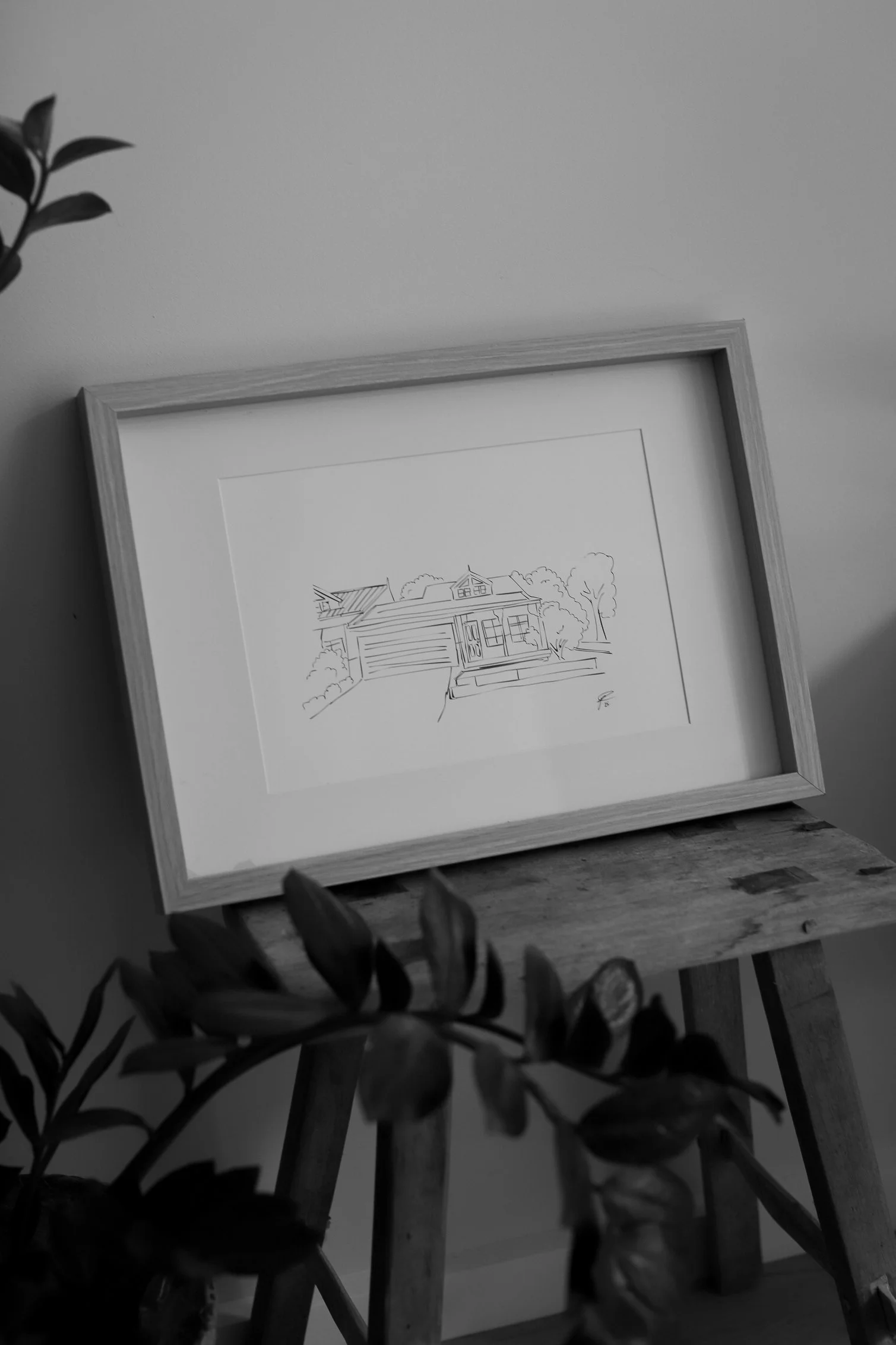 Black and white photo of a framed house sketch placed on a small wooden table, with leafy plants in the foreground and a plain wall background.