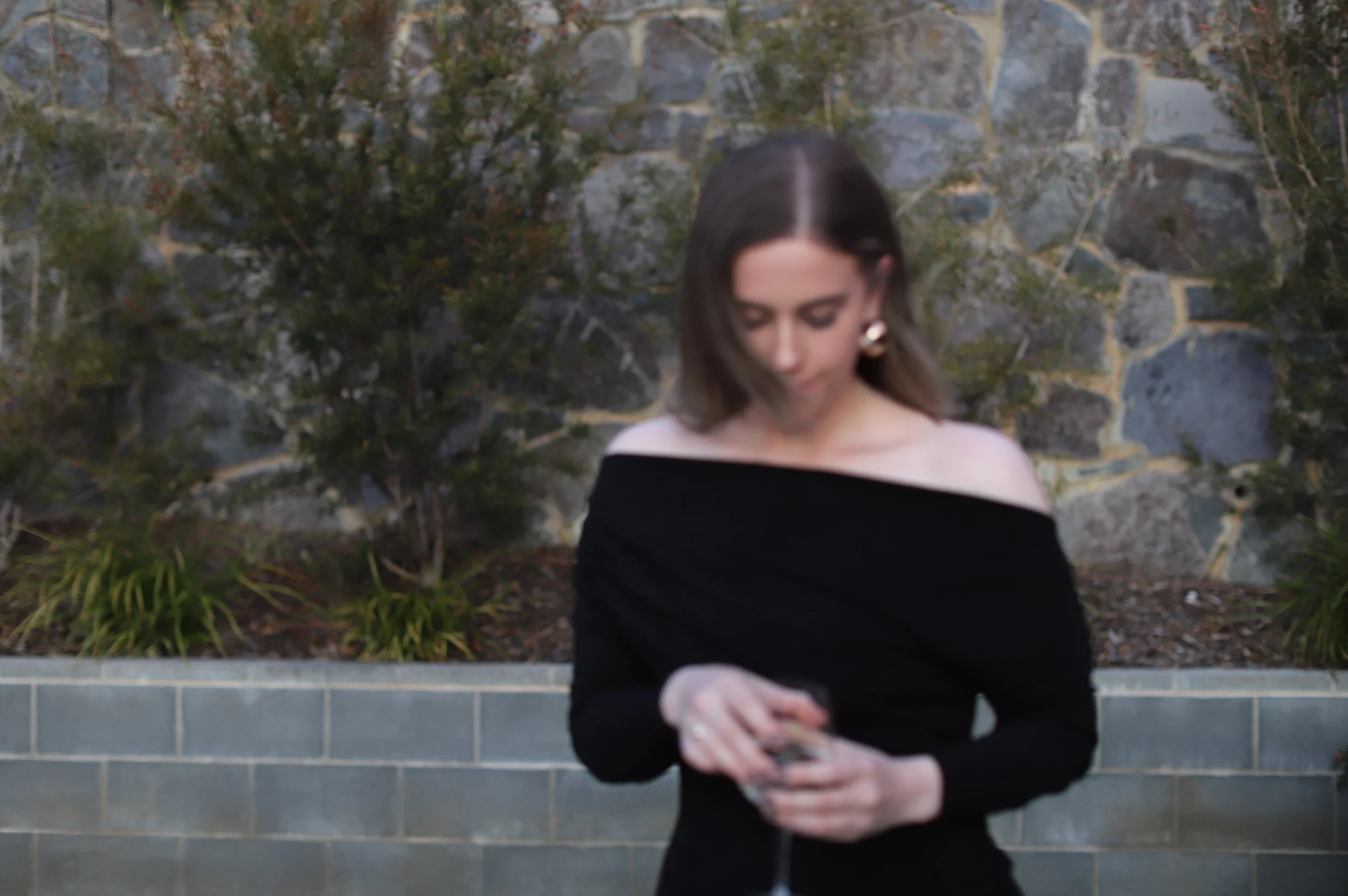 A woman looking down at her phone outdoors, wearing an off-the-shoulder black dress, with a stone wall and greenery in the background.