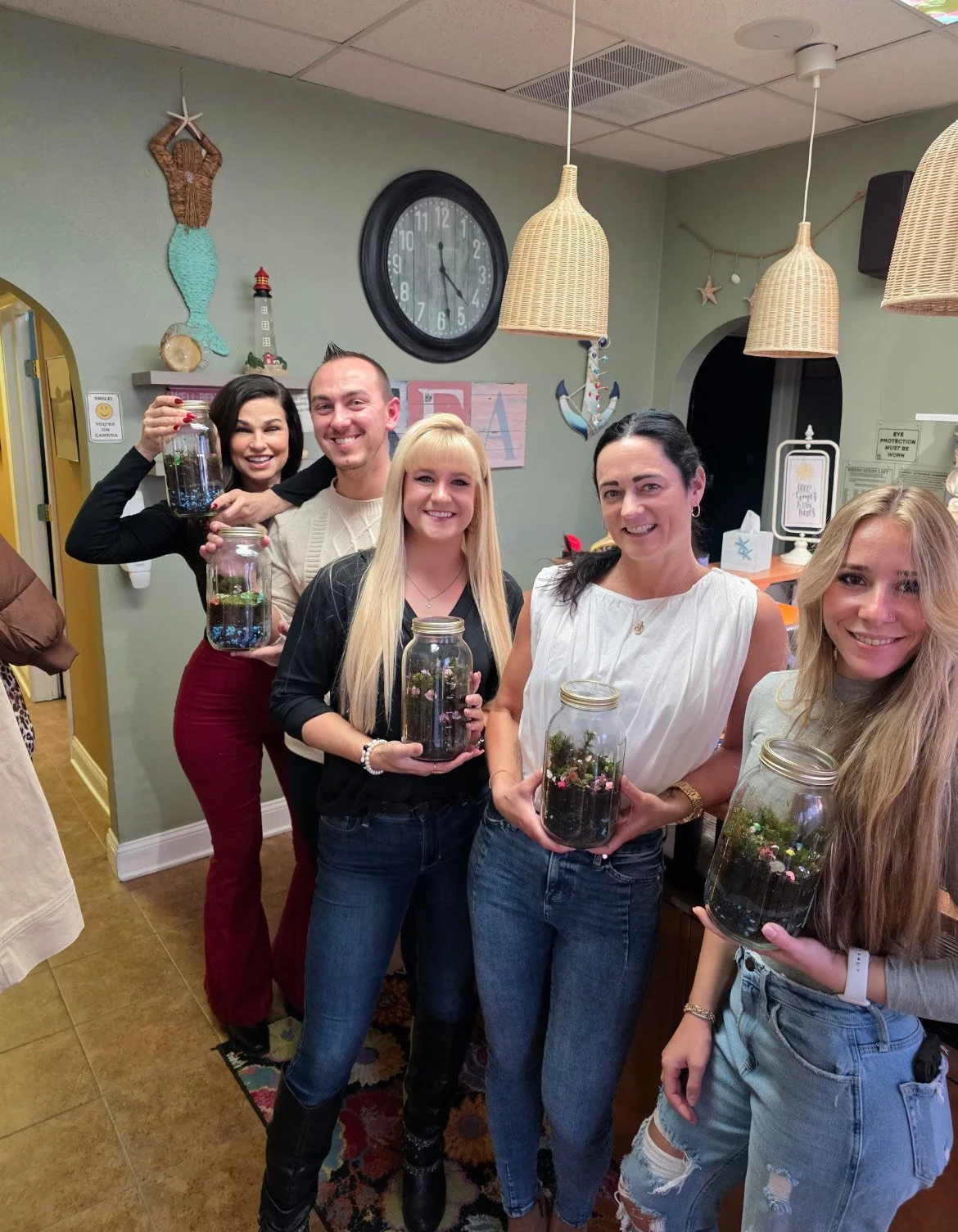 Group of six women and one man holding glass jars with plants inside at a party or gathering indoors, with decorations on the wall and hanging lamps above.