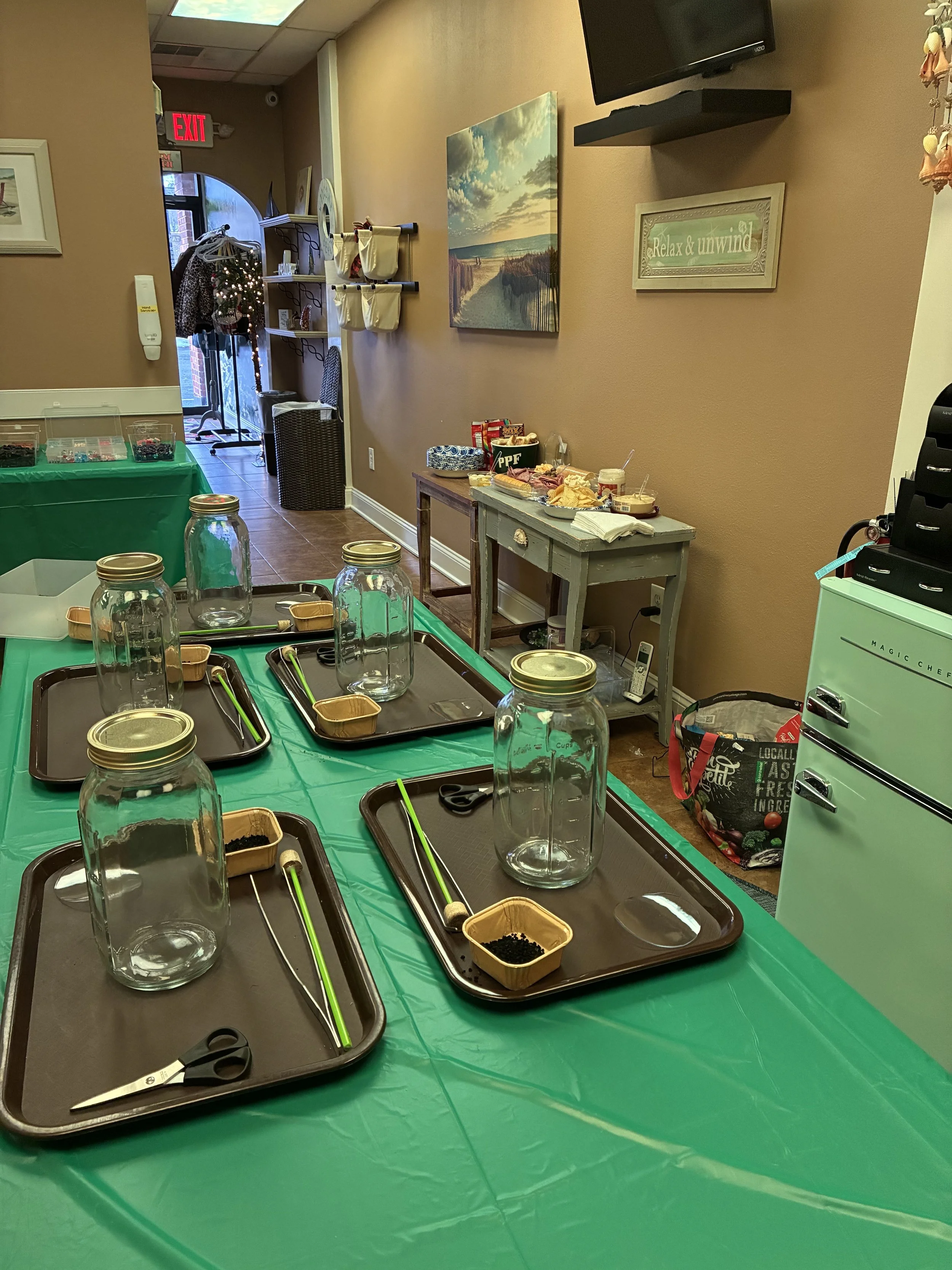 Table with four glass jars and small containers, likely for a craft or DIY activity, in a room with a decorated wall, a small table with snacks, and a nearby food and beverage station.