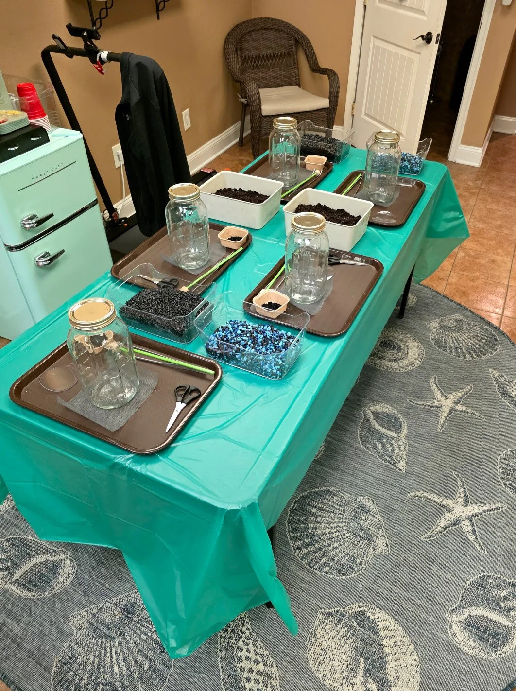 Table with craft supplies, jars, small containers of black and blue beads, scissors, and trays, set up for a craft activity. The table is covered with a green tablecloth, and a wicker chair and a mini refrigerator are in the background.