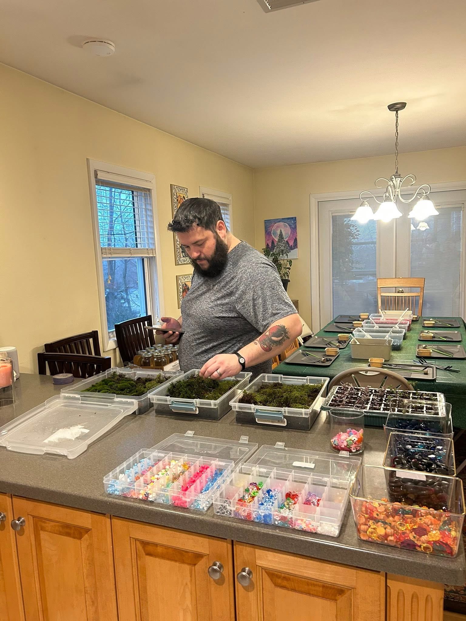 Man with a beard and tattoo on his arm organizing jewelry or craft supplies on a kitchen island, with craft supplies, jewelry trays, and containers of beads and decorations on the counter.