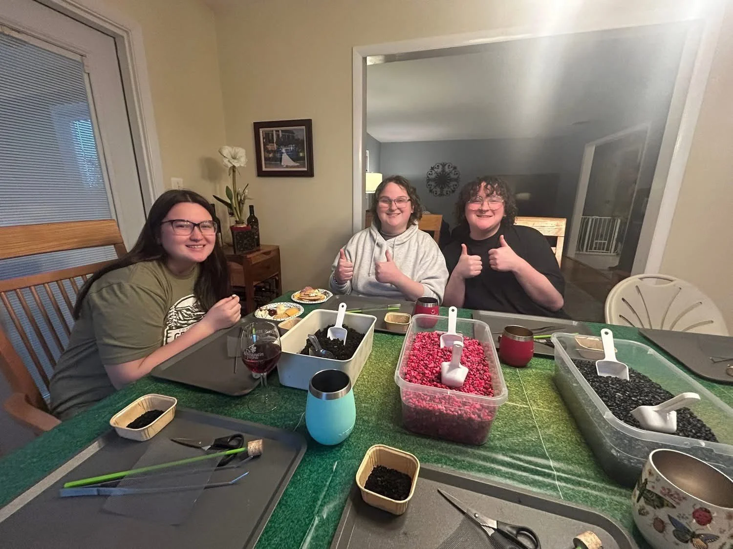Three smiling people seated at a dining table with bowls of berries and utensils, giving thumbs up.