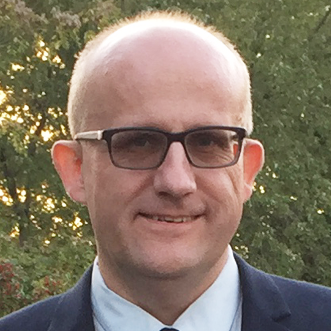 A middle-aged man with glasses, short light hair, wearing a suit and tie, standing outdoors with trees in the background.