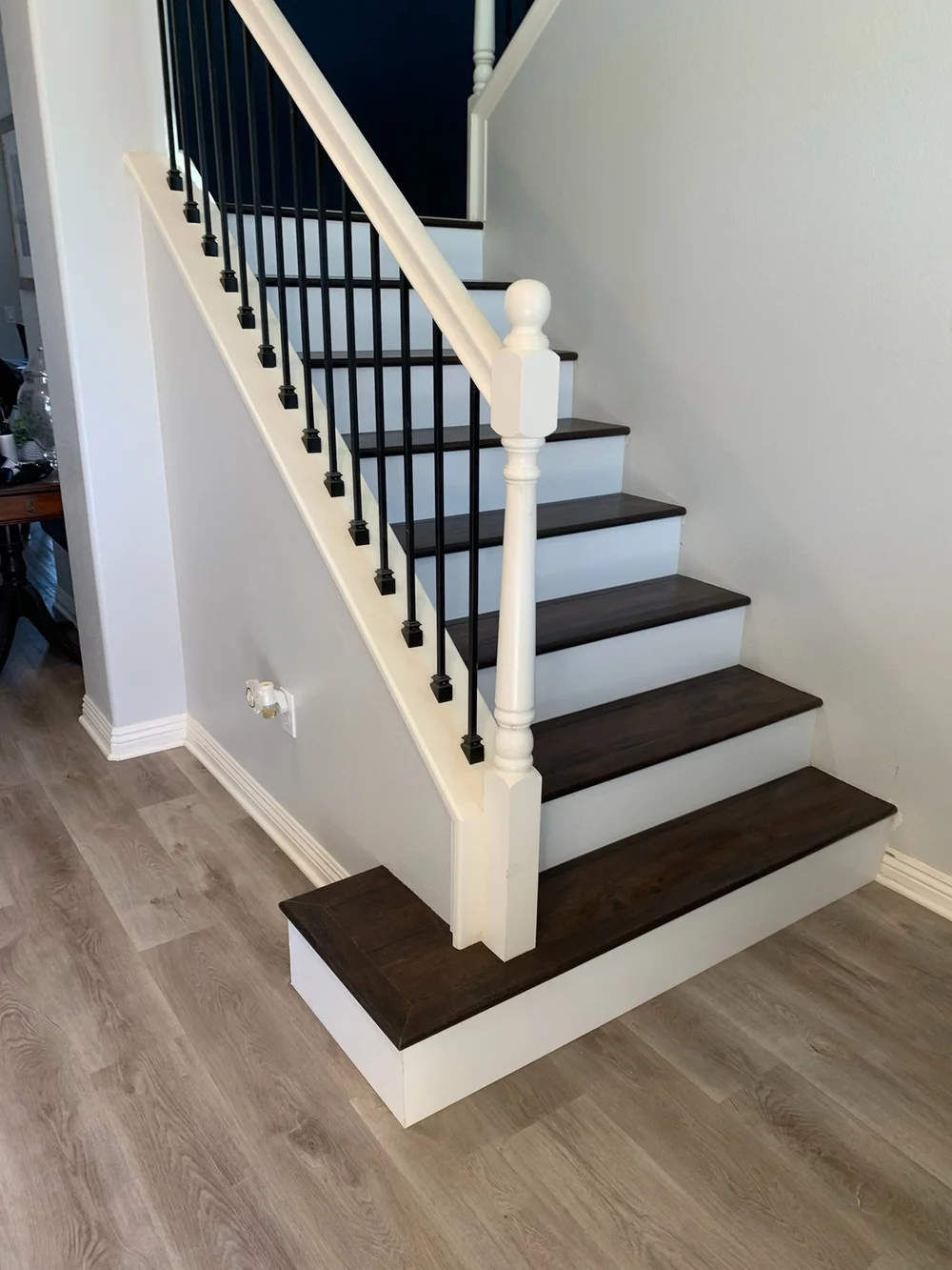 Stair remodel with dark-stained wood treads, white risers and black iron balusters beside wood-look plank flooring