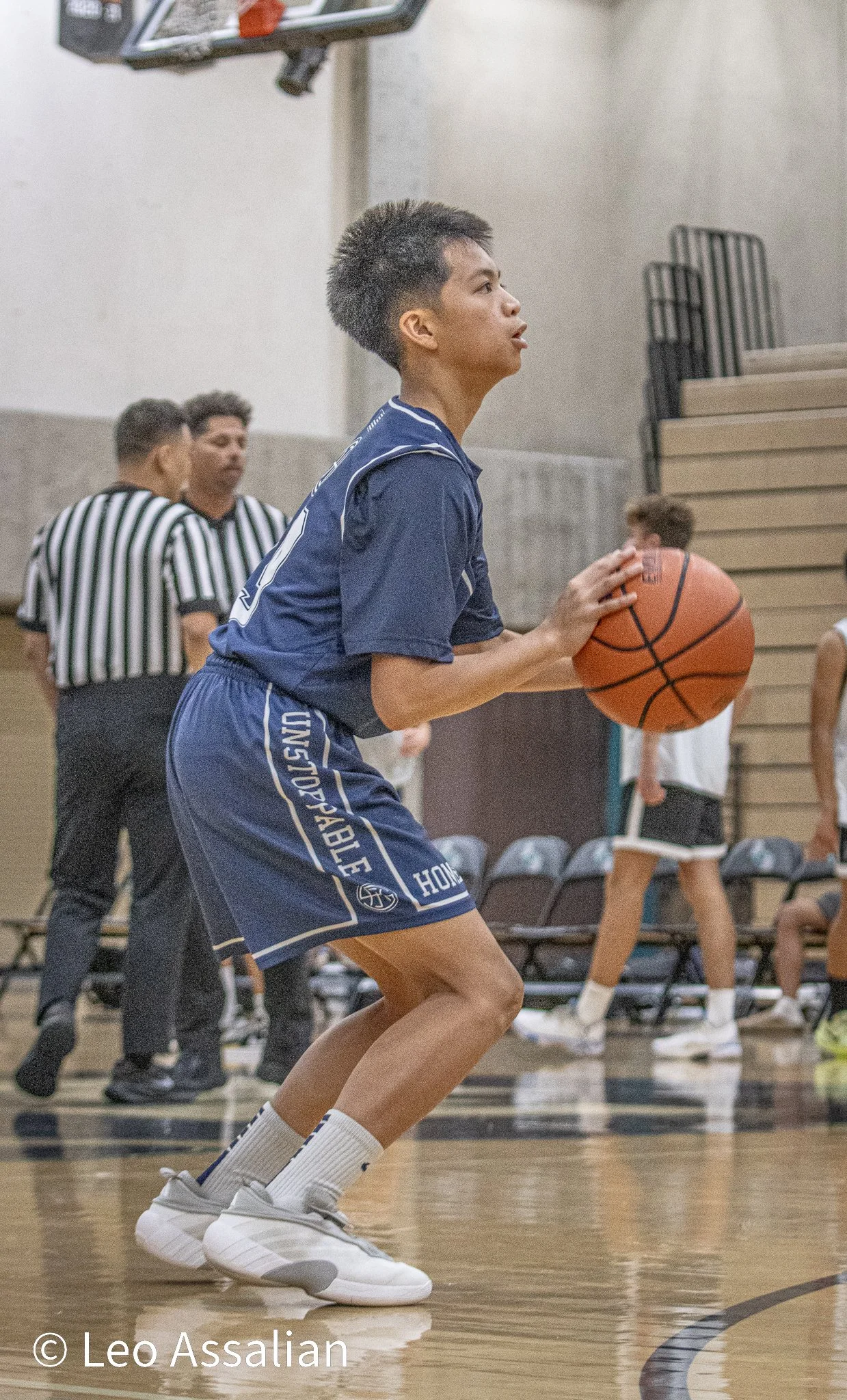 A young male basketball player in a blue uniform ready to shoot a basketball on an indoor court, with referees and other players in the background.