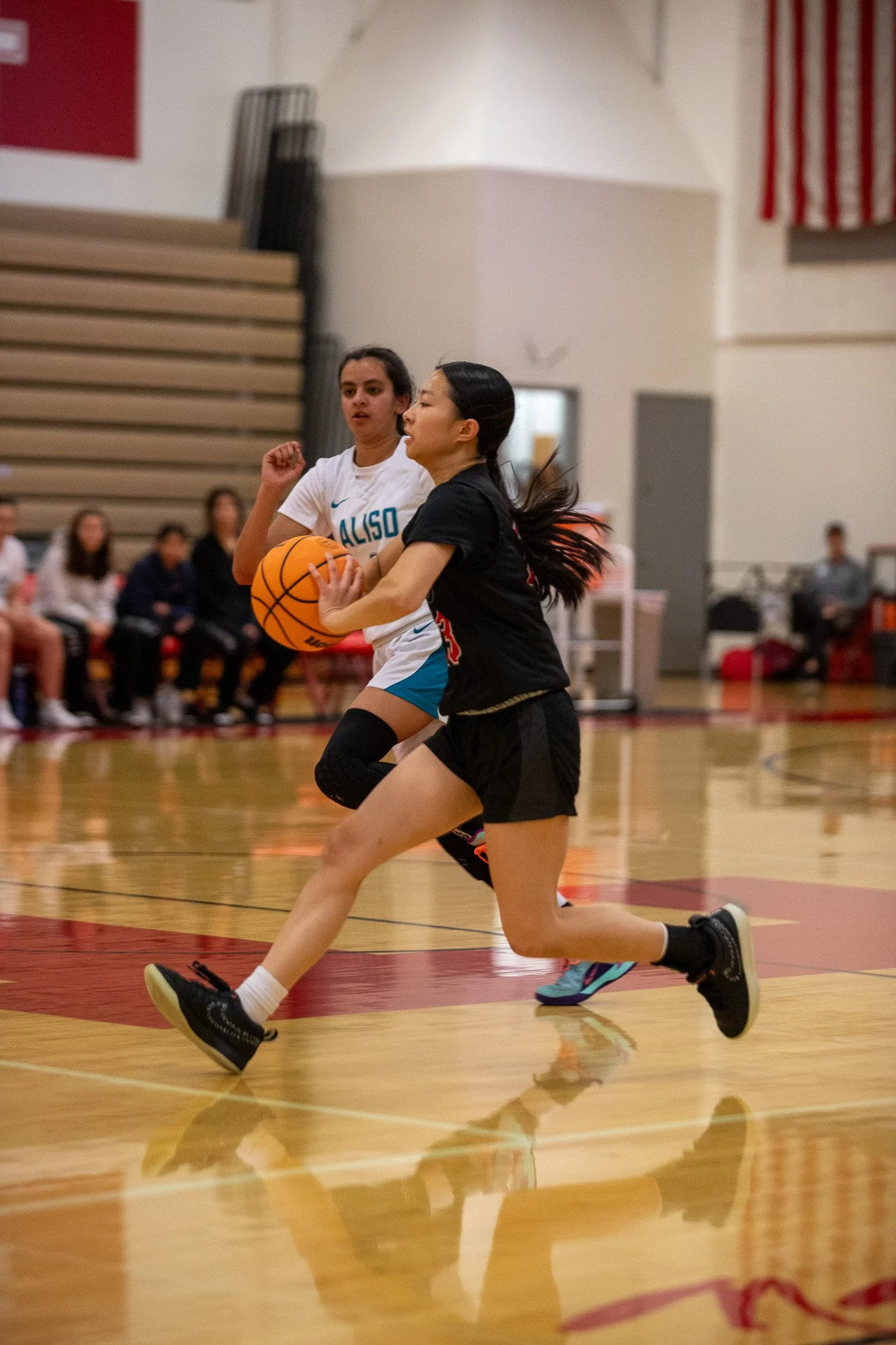 Two girls playing basketball on an indoor court, one in a white jersey and the other in a black jersey, with spectators watching from the bench in the background.