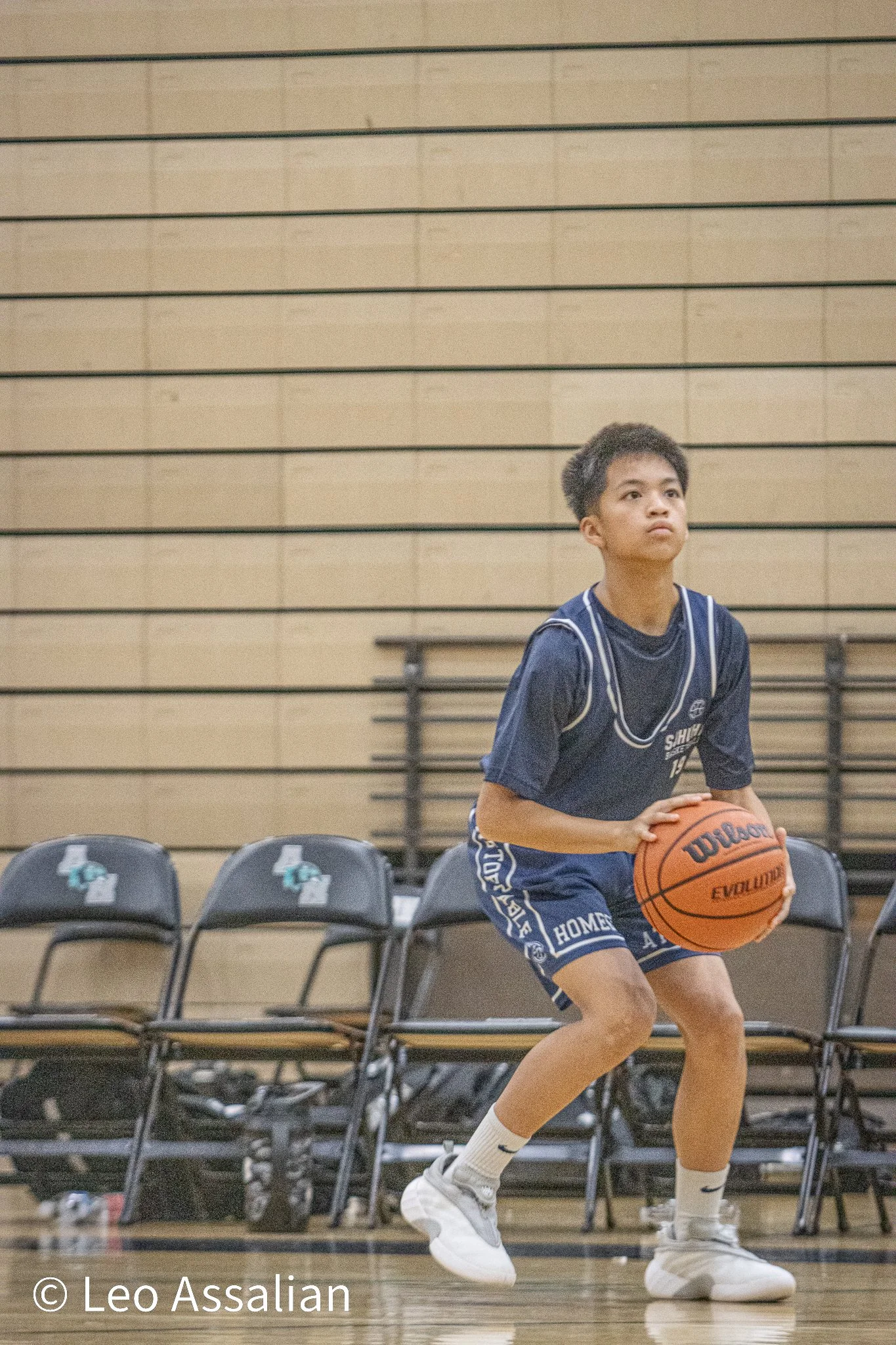A young boy in a navy basketball uniform practicing free throws in a gymnasium, holding an orange Wilson basketball and standing on a wooden court near black chairs with logos.