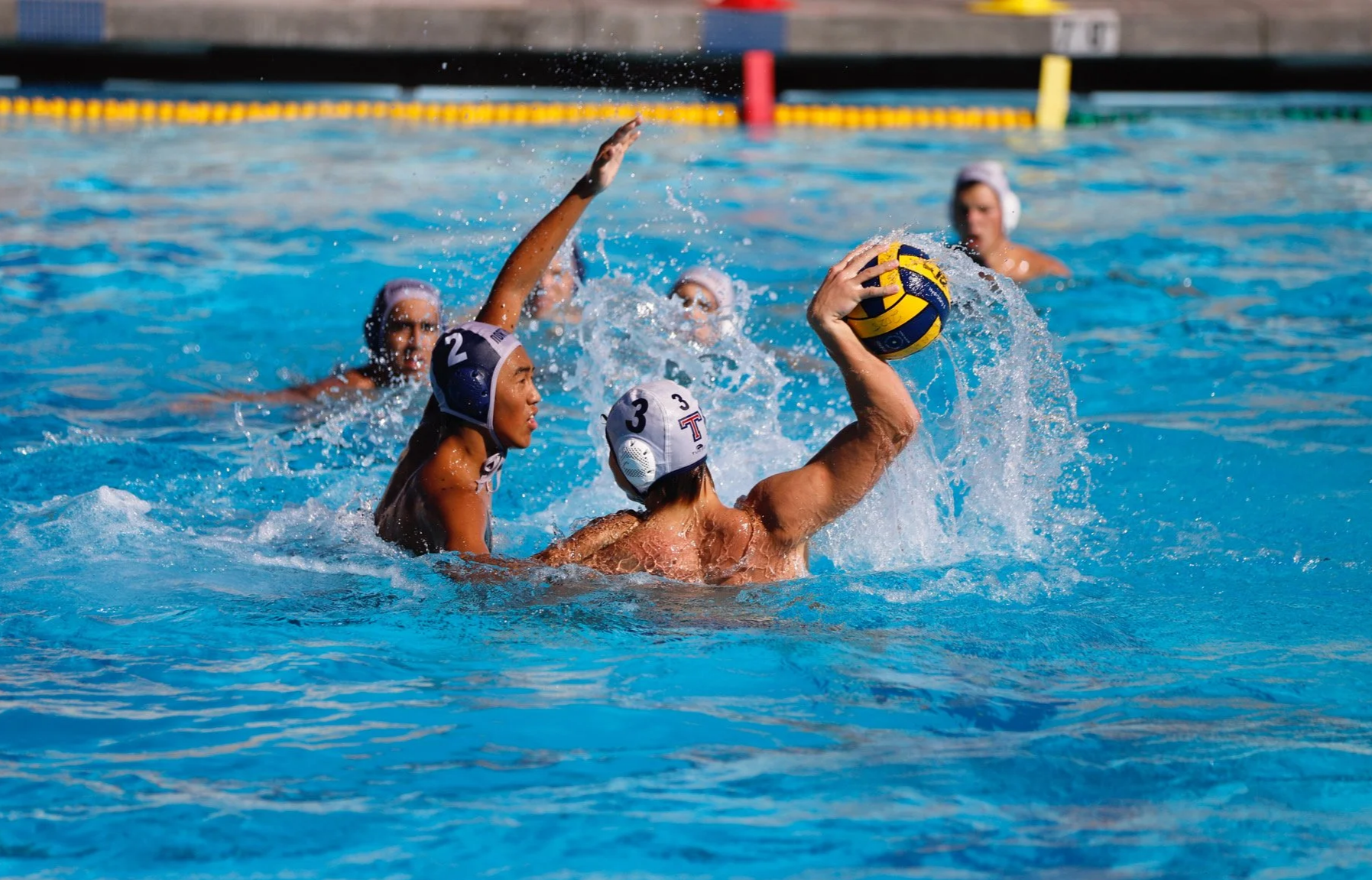 Water polo players in action during a game, with one player holding the ball above the water and others defending, all wearing caps and goggles in a swimming pool.