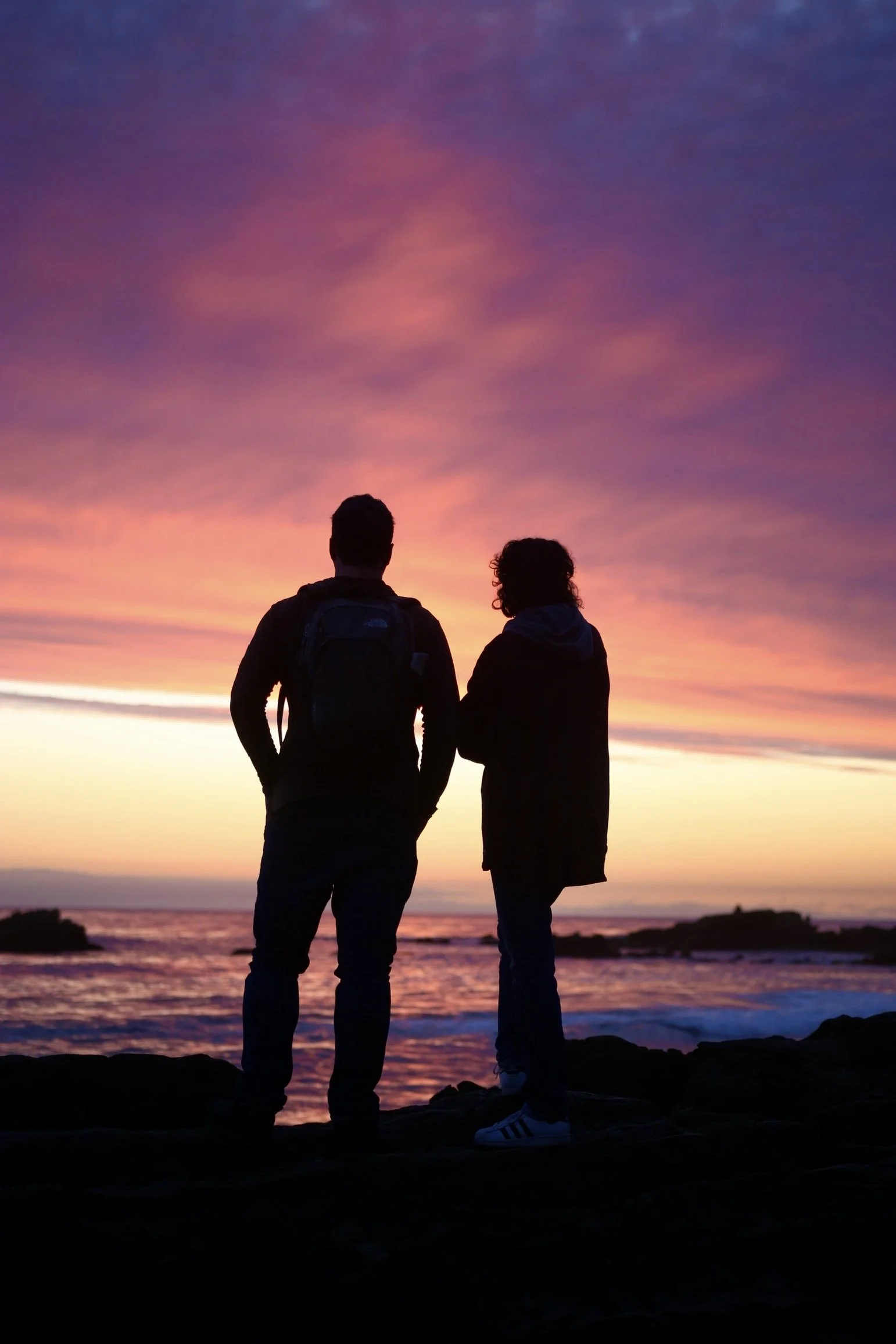 Silhouettes of a man and woman standing on rocks at the beach during sunset, with colorful pink, purple, and orange sky.